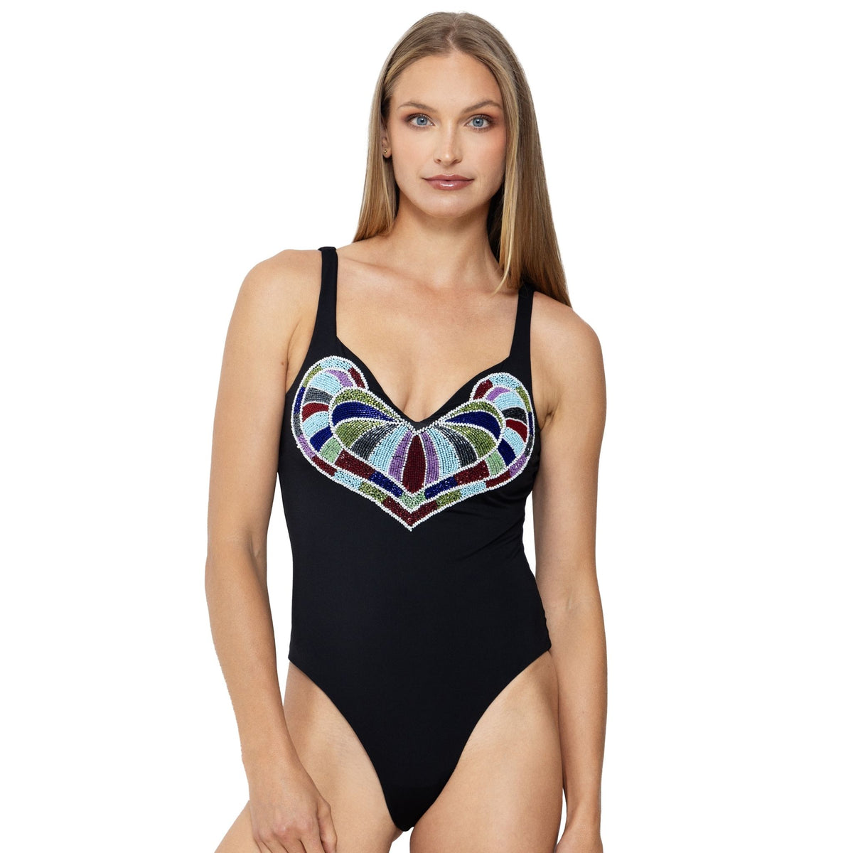 A woman with long blonde hair models the SELF-ADORE Amor Hand-Beaded Embellished One-Piece Swimsuit in black, featuring a colorful geometric heart design on the chest. She stands against a white background, looking at the camera.