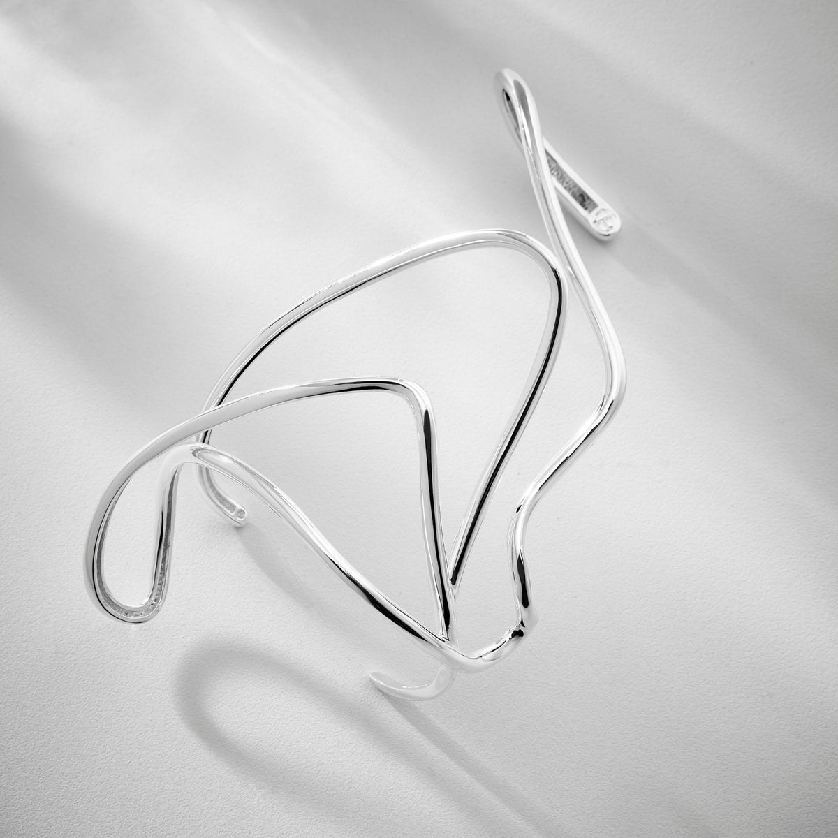 The Silver Miragem Hand Bracelet from ANNA PRATA’s Duna Collection features fluid, sculptural curves with elegant gold and silver plating, casting artistic shadows as it rests on a white surface.