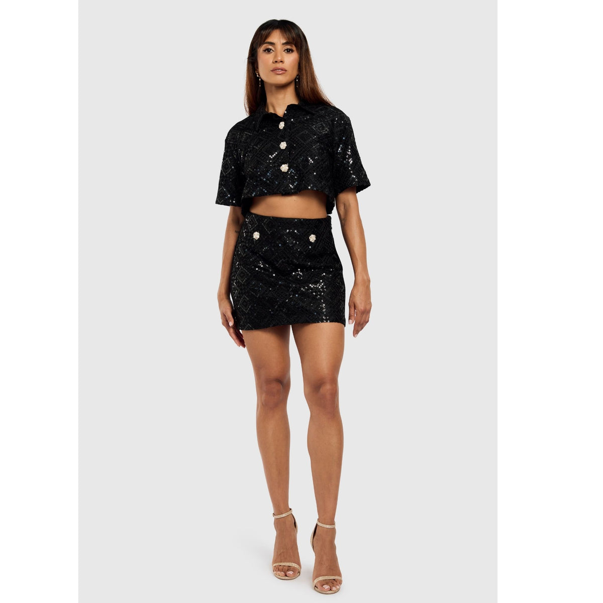A woman faces forward in a black INTERNATIONAL APPEAL Sequin Mini Skirt | PR Sample with a matching sequin crop top, large buttons, and high-heeled sandals. She has long hair and poses against a plain light gray background—ideal for evening wear.