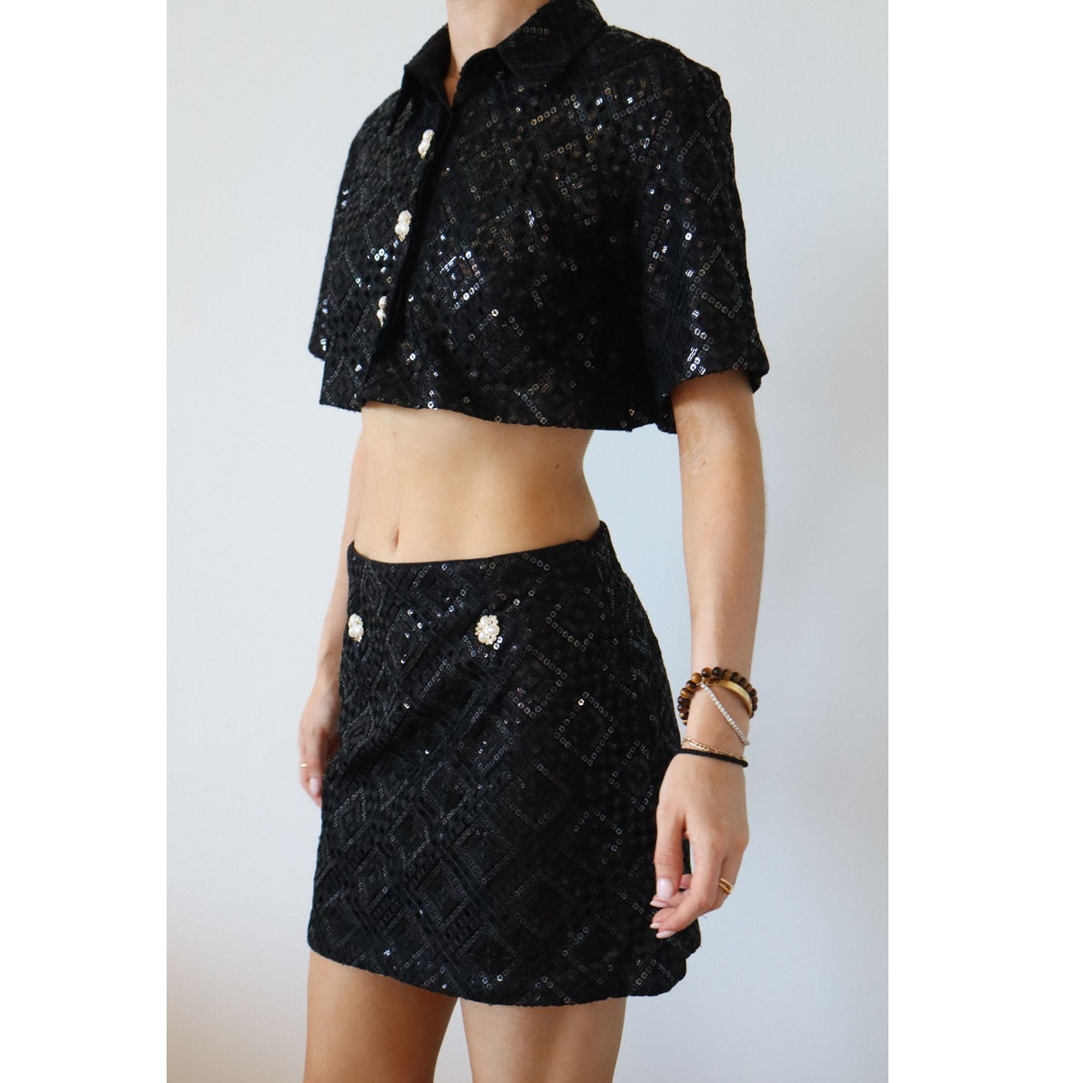 A woman models the INTERNATIONAL APPEAL Sequin Co-Ord Crop Shirt | PR Sample, a black short-sleeve sequin crop shirt and high-waisted mini skirt with decorative buttons. She accessorizes with bracelets and rings; her head is not visible.