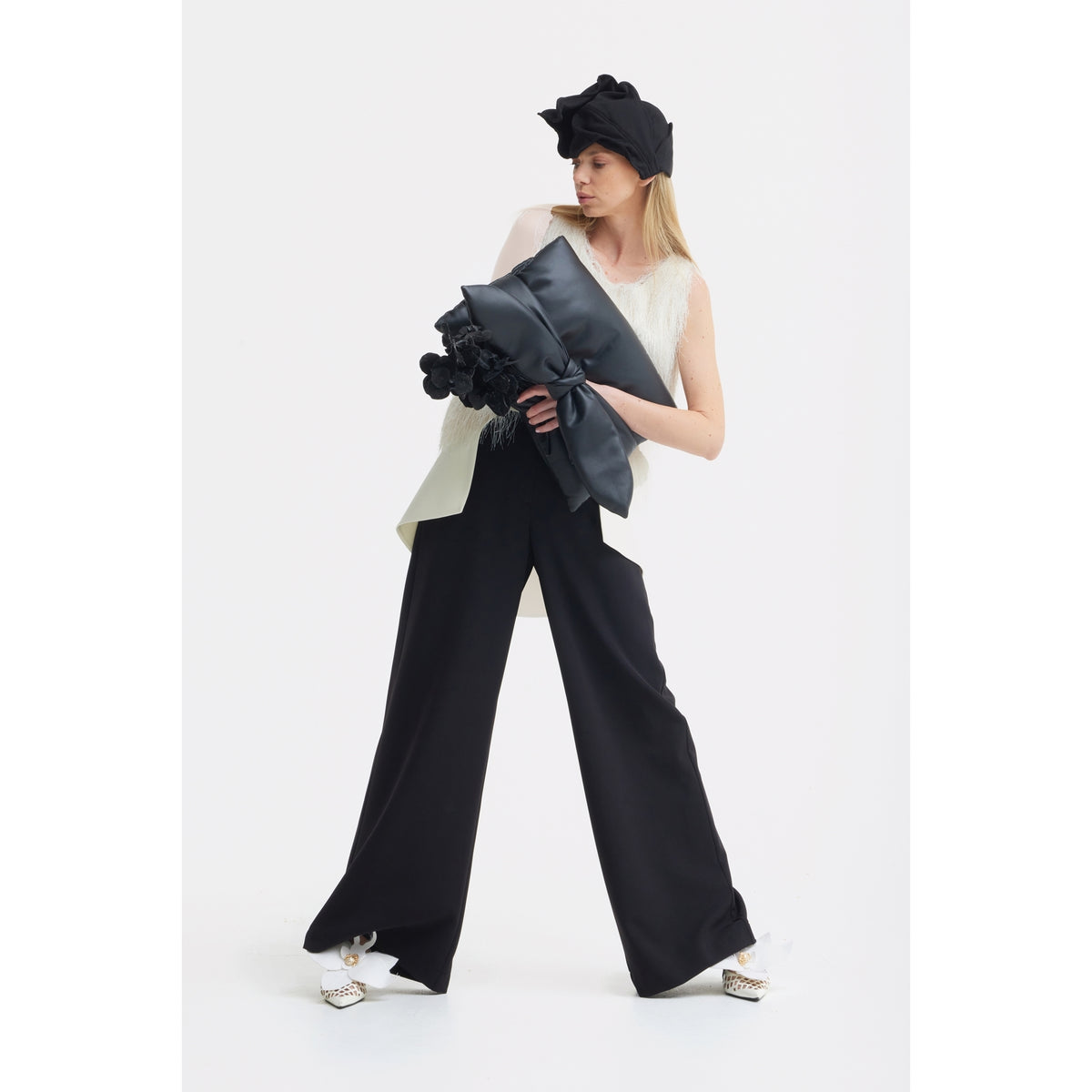 A woman poses against a white background in a sleeveless white top, JULIA ALLERT Statement Waist Wide-Leg Trousers in black, a large black bow hat, white pointed shoes, and holding a large black bow with floral details.