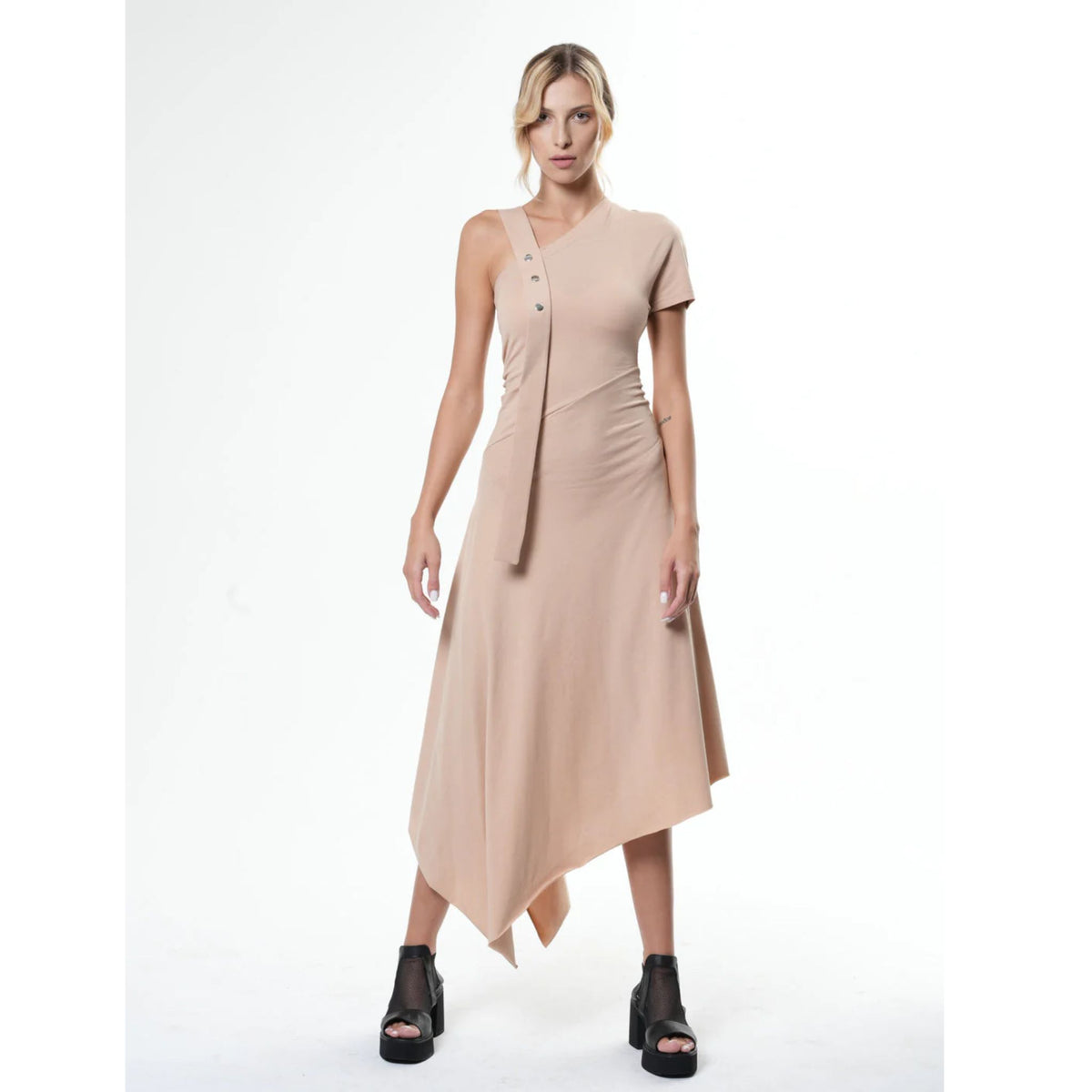 A woman faces forward in the METAMORPHOZA Asymmetric Cotton Beige Dress (PR Sample), featuring a handkerchief hem and decorative strap with buttons. She wears her blonde hair in a loose ponytail and pairs the look with black platform sandals.