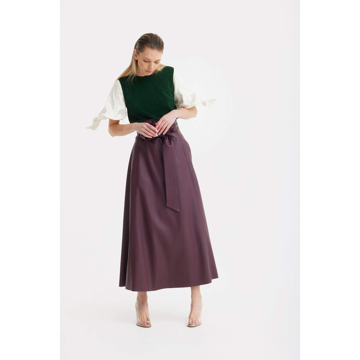 A woman wearing the JULIA ALLERT Green Lurex Knit Top With Adjustable Satin Sleeves, paired with a high-waisted, ankle-length purple skirt and clear heeled shoes, stands against a white background.