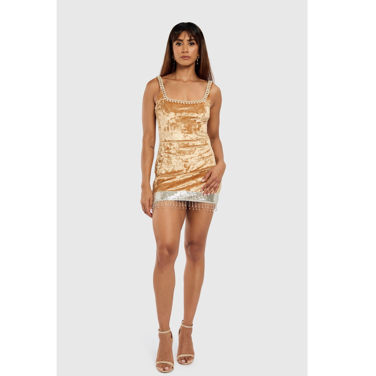 A woman with straight brown hair and a neutral expression models the INTERNATIONAL APPEAL Mustard Velvet Pearl Mini Dress | PR Sample, featuring hand-stitched pearl trim and rhinestone straps, paired with nude heels. Made in India.