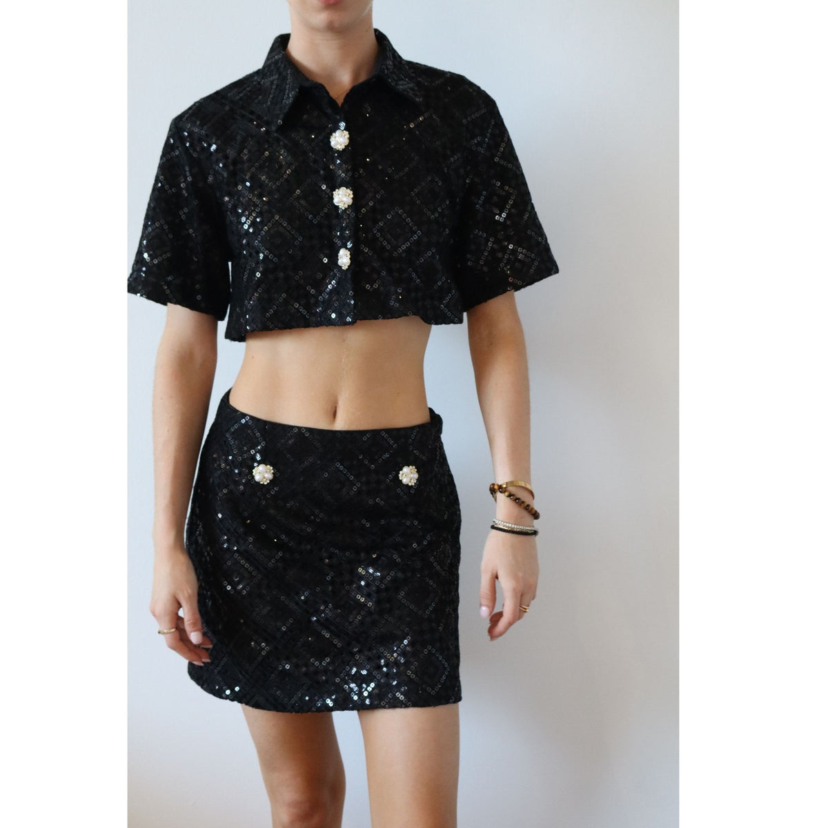 A person wears the INTERNATIONAL APPEAL Sequin Mini Skirt | PR Sample, a limited edition black checkered sequin mini skirt styled with a cropped short-sleeve top featuring decorative buttons—ideal for chic evening wear.