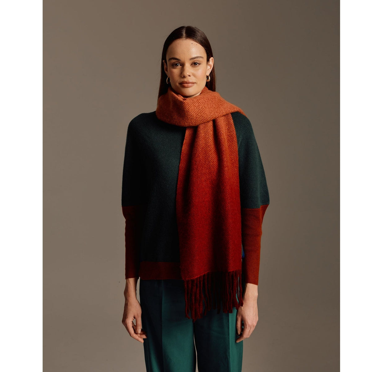 A woman with straight brown hair wears a dark green sweater, teal pants, and the OO-TO Standard length Fringed Ombre Scarf in wine and rust, standing against a plain brown background.