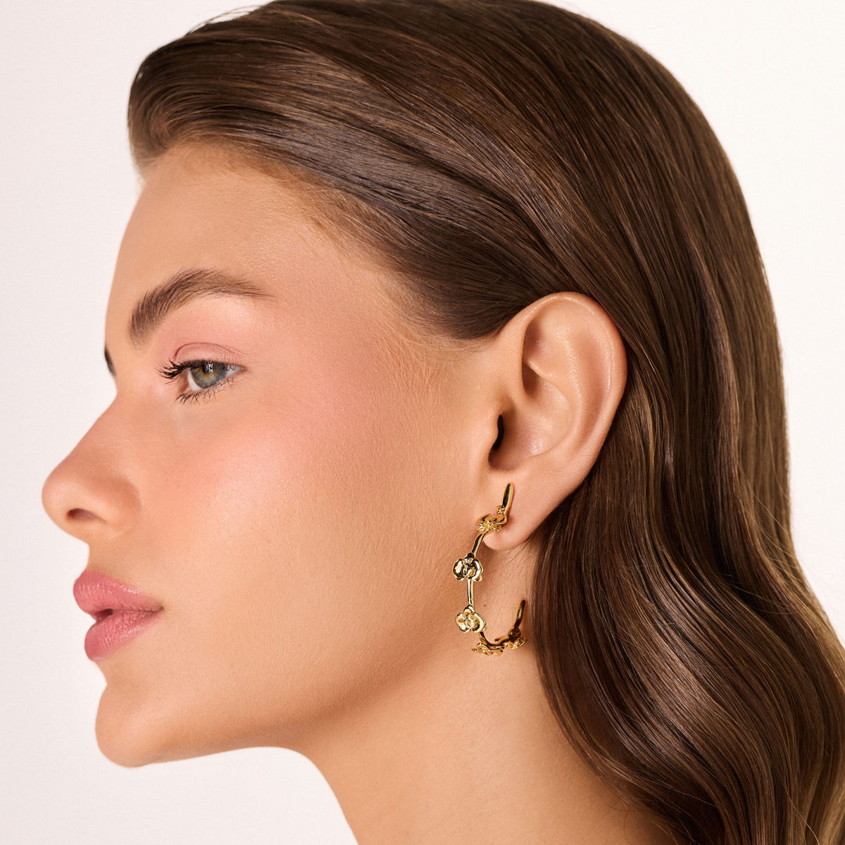 A woman with smooth, brown, wavy hair is shown in profile wearing large Gold Floralis Hoops from the AFlora Collection by ANNA PRATA. She has natural makeup with soft pink eyeshadow and lipstick, set against a plain white background.