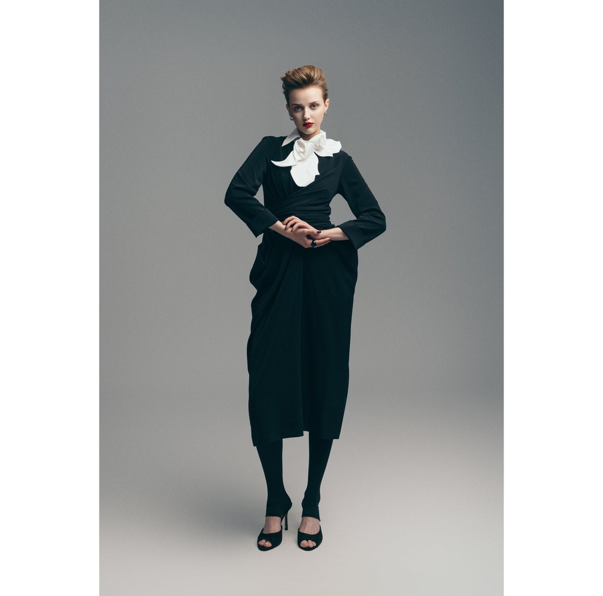 A woman models the JULIA ALLERT Designer Neckwear - Collar in white—an oversized satin bow at the neck—paired with a black long-sleeve dress, black tights, and open-toed heels against a plain gray background.