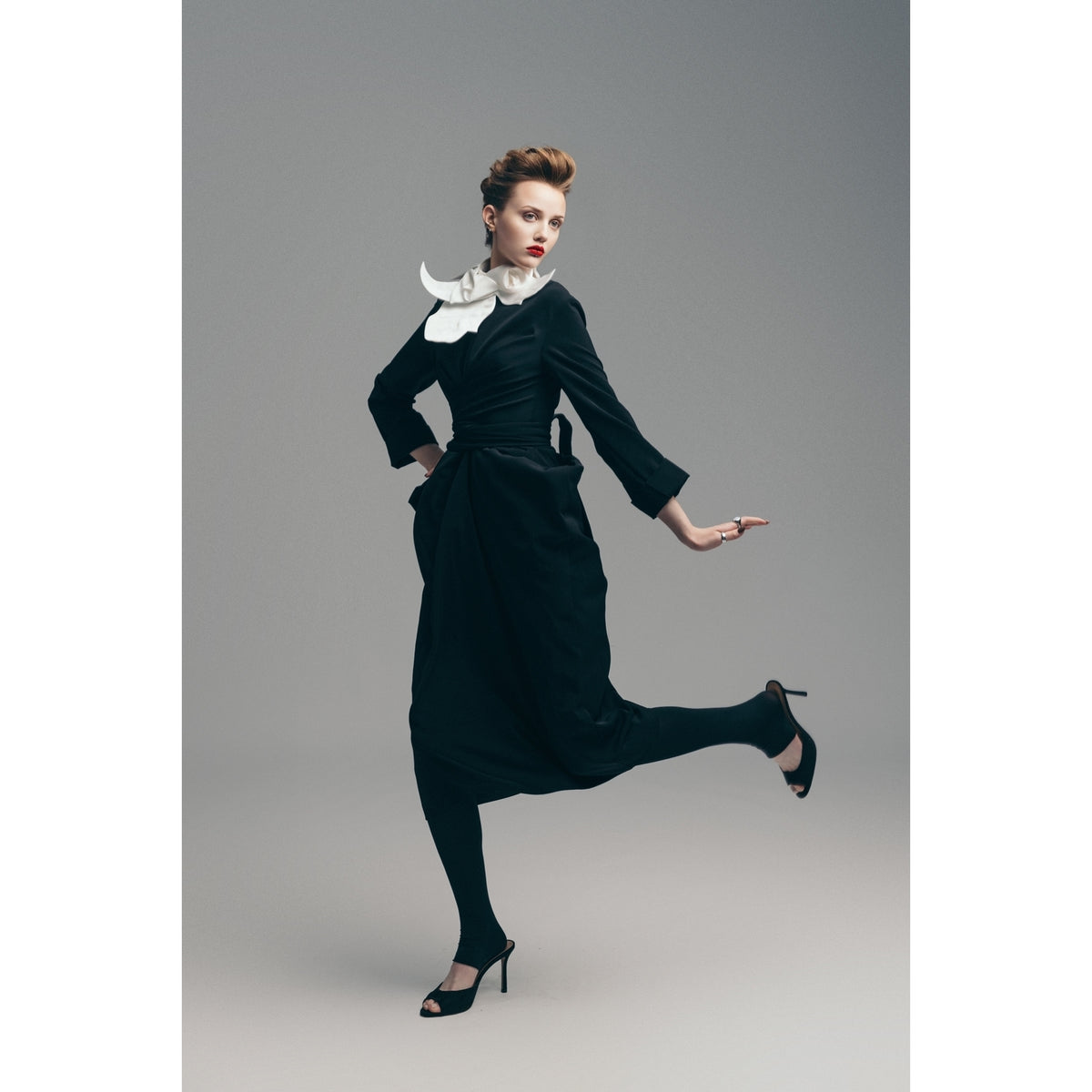 A woman strikes a pose in a chic black dress, featuring the JULIA ALLERT Designer Neckwear - Collar in white, paired with black tights and high heels, set against a plain gray background.