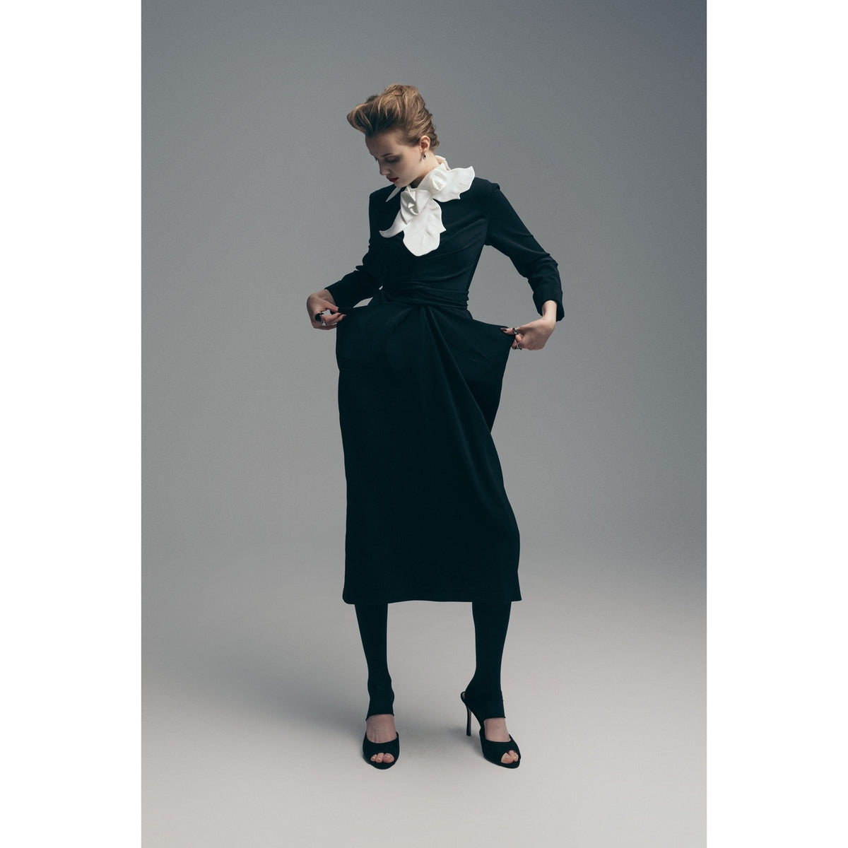 A person wears the JULIA ALLERT Designer Neckwear - Collar in white with a long-sleeved black dress, black open-toe heels, and leggings, standing against a plain background and looking down while holding their dress.