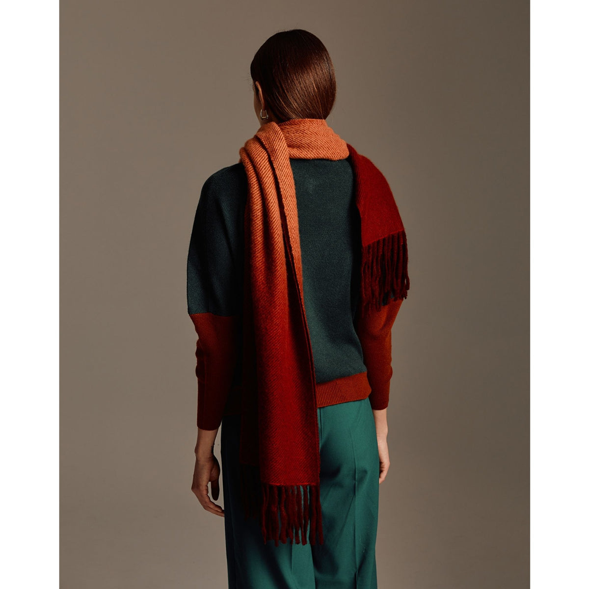 A person with straight brown hair, seen from behind, wears a dark green and rust color-blocked sweater, teal pants, and an OO-TO standard length fringed ombre scarf in wine and rust. The background is plain and neutral.