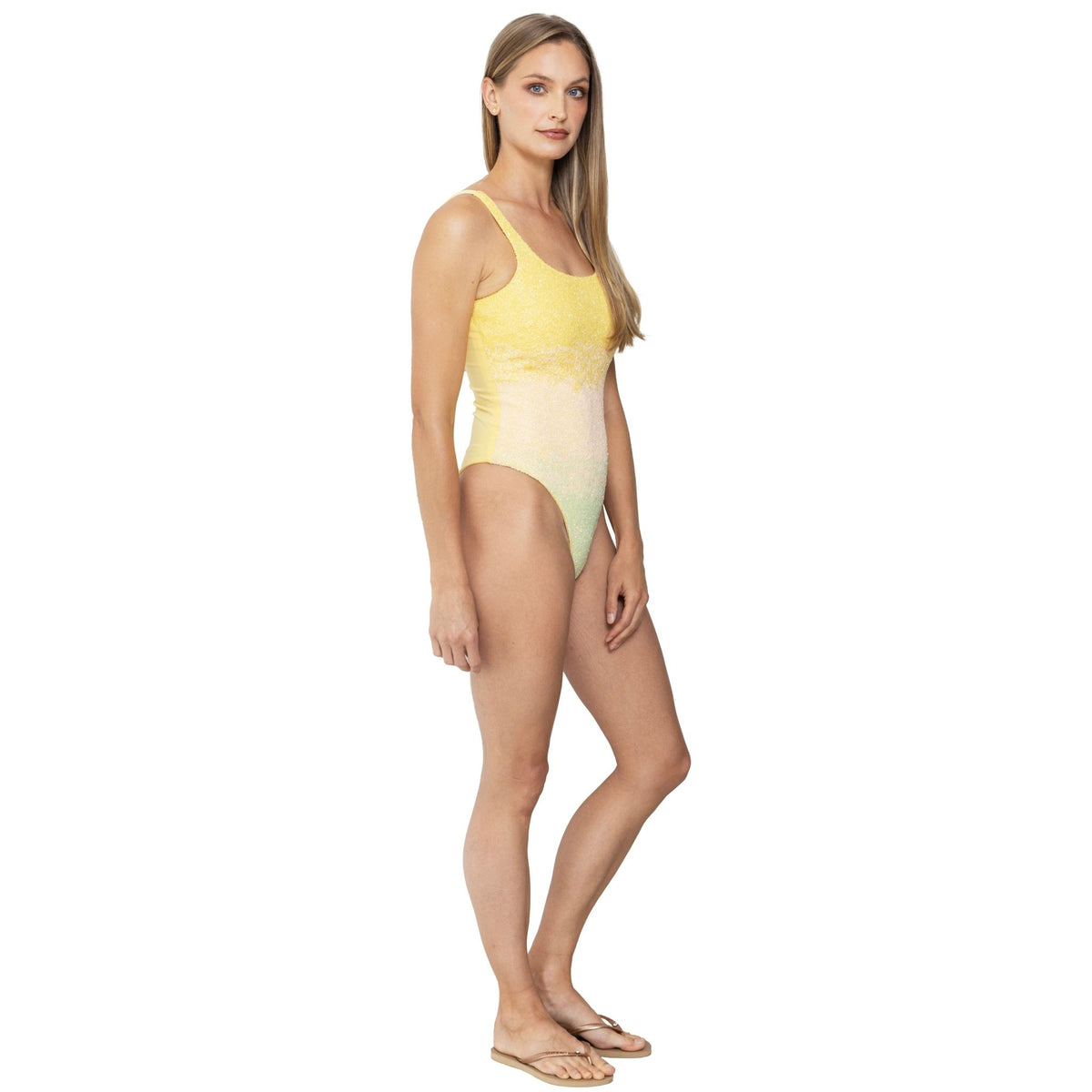 A woman with long blonde hair wears the SELF-ADORE Ombre Hand-Beaded Embellished One-Piece Swimsuit in yellow, paired with beige sandals, standing against a white background with a slight side angle and neutral expression.