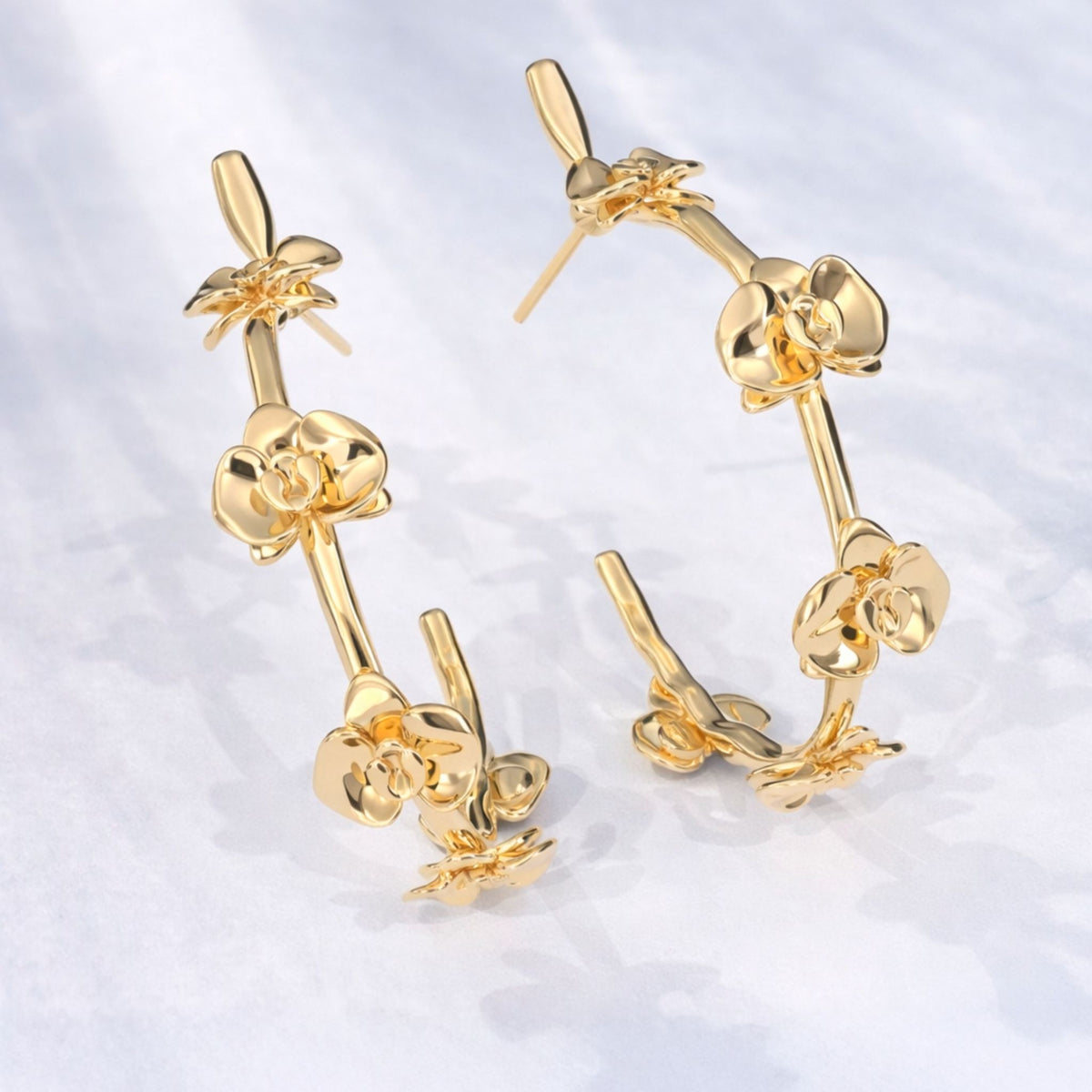 Two Gold Floralis Hoops from the AFlora Collection by ANNA PRATA, featuring delicate flower-inspired designs, are displayed on a light textured surface with soft shadows.