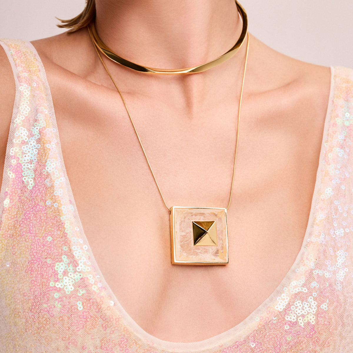 A close-up of a person wearing a sequined light pink top and two gold necklaces—a thick choker and the Gold Vértice L Necklace from ANNA PRATA's Faces Collection, featuring a square pendant with a central triangle design.