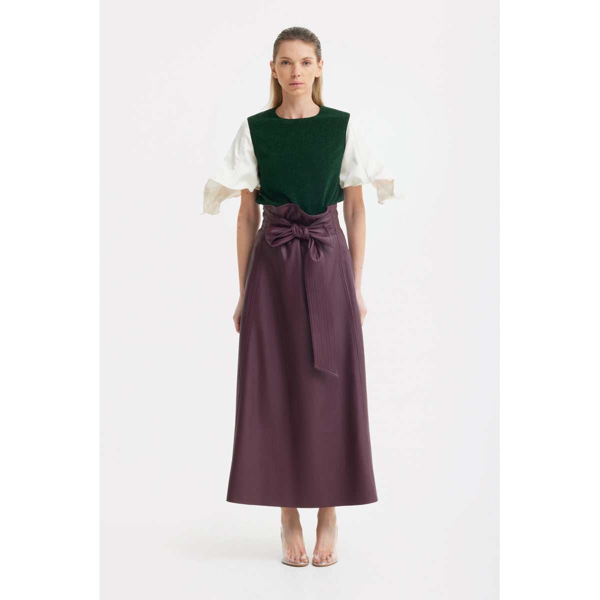 A woman with long straight hair stands against a plain background, wearing a dark green top with white ruffled sleeves and the JULIA ALLERT Faux Leather High-Waisted A-Line Skirt With Belt in Burgundy, styled with heels.