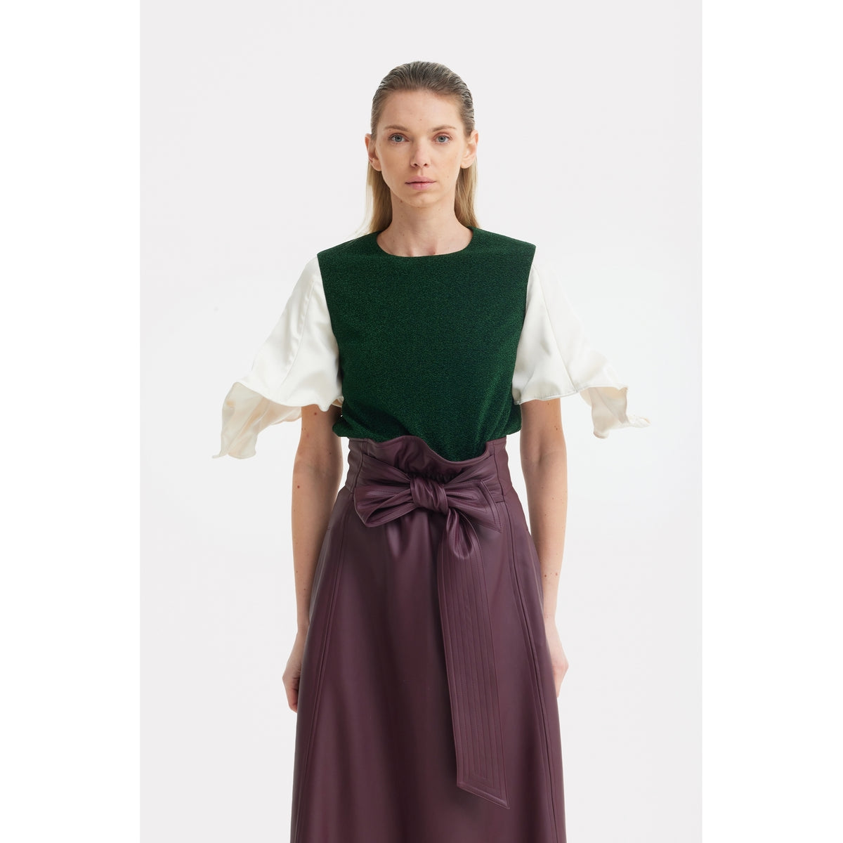 A woman with straight light brown hair and a neutral expression wears a green top with short, ruffled white sleeves and the JULIA ALLERT Faux Leather High-Waisted A-Line Skirt With Belt in Burgundy.