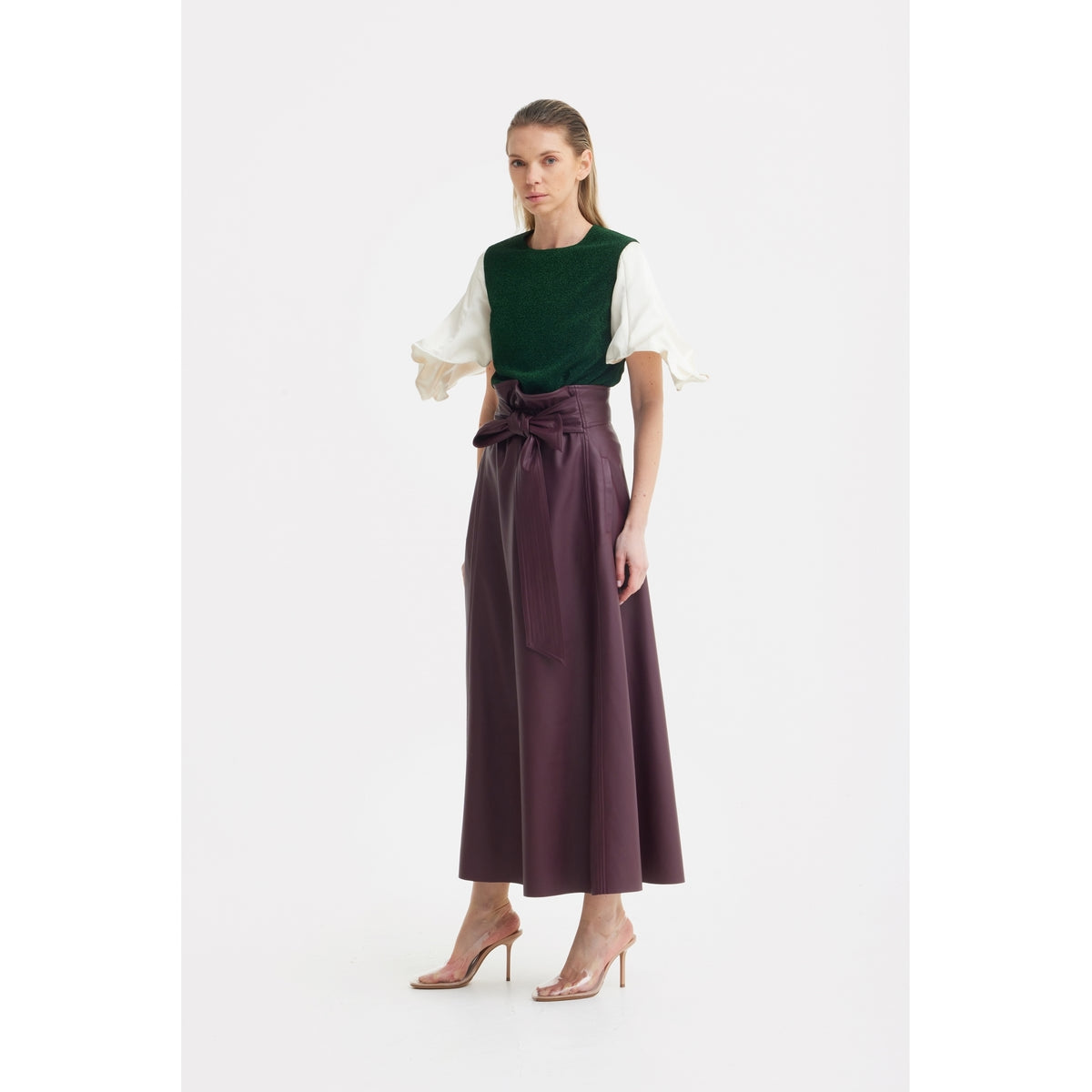 A woman faces forward in a green top with white puff sleeves, styled with the JULIA ALLERT Faux Leather High-Waisted A-Line Skirt With Belt in Burgundy. She wears clear high-heeled sandals and has long hair, set against a white background.