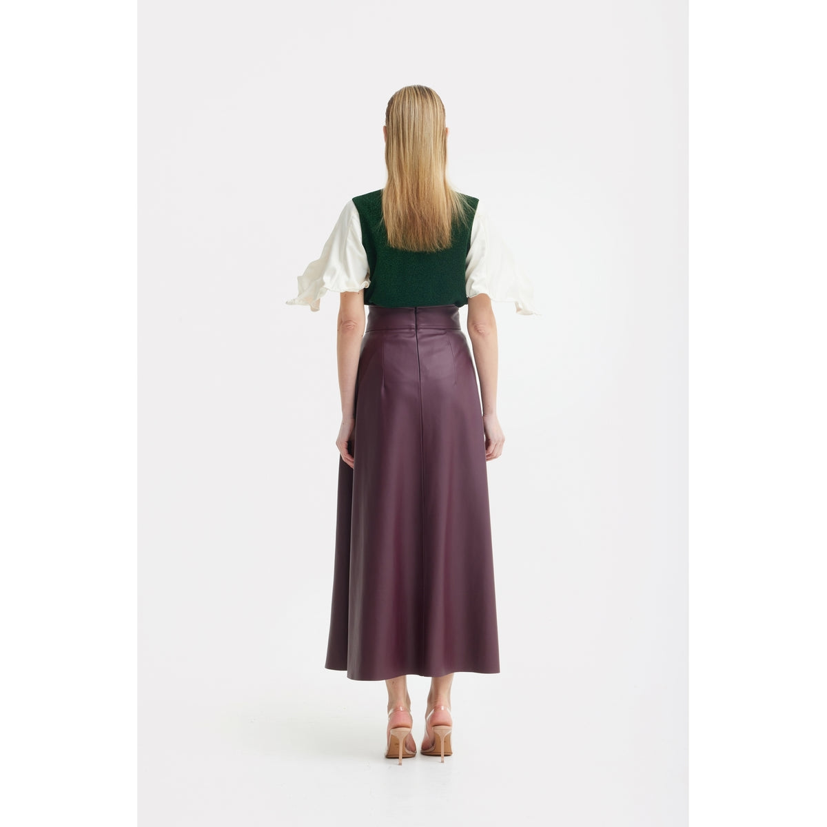 A woman stands facing away against a plain white background, wearing a dark green vest, white puffy-sleeve blouse, nude heels, and the JULIA ALLERT Faux Leather High-Waisted A-Line Skirt With Belt in Burgundy.