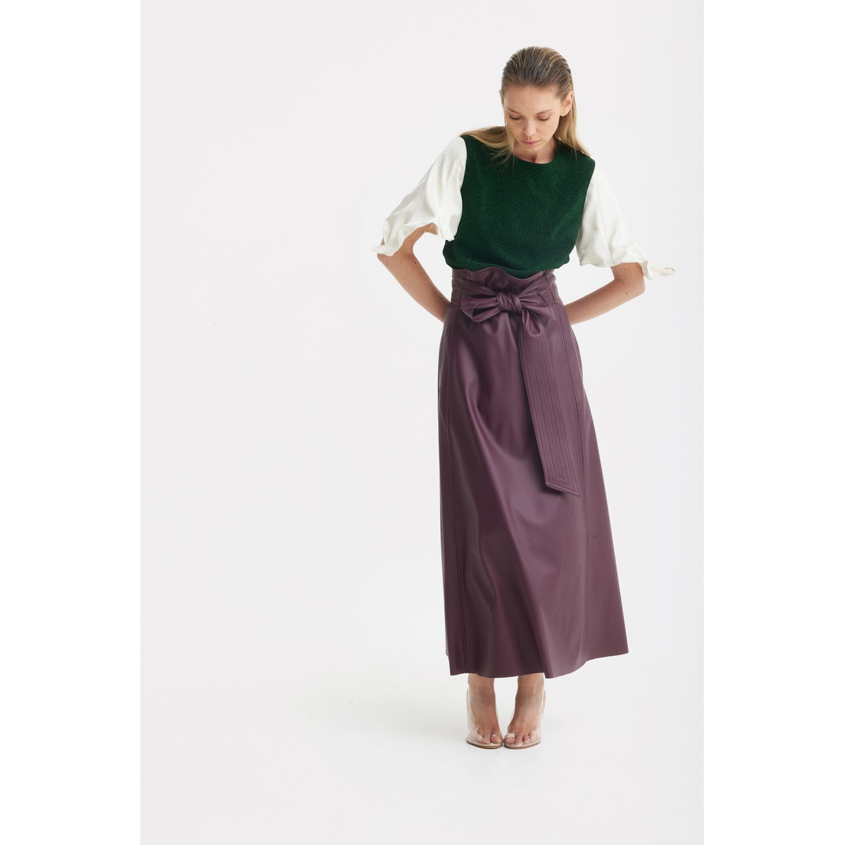 A woman stands against a white background in a dark green top and a JULIA ALLERT Faux Leather High-Waisted A-Line Skirt With Belt in burgundy, looking down with her hands on her hips.