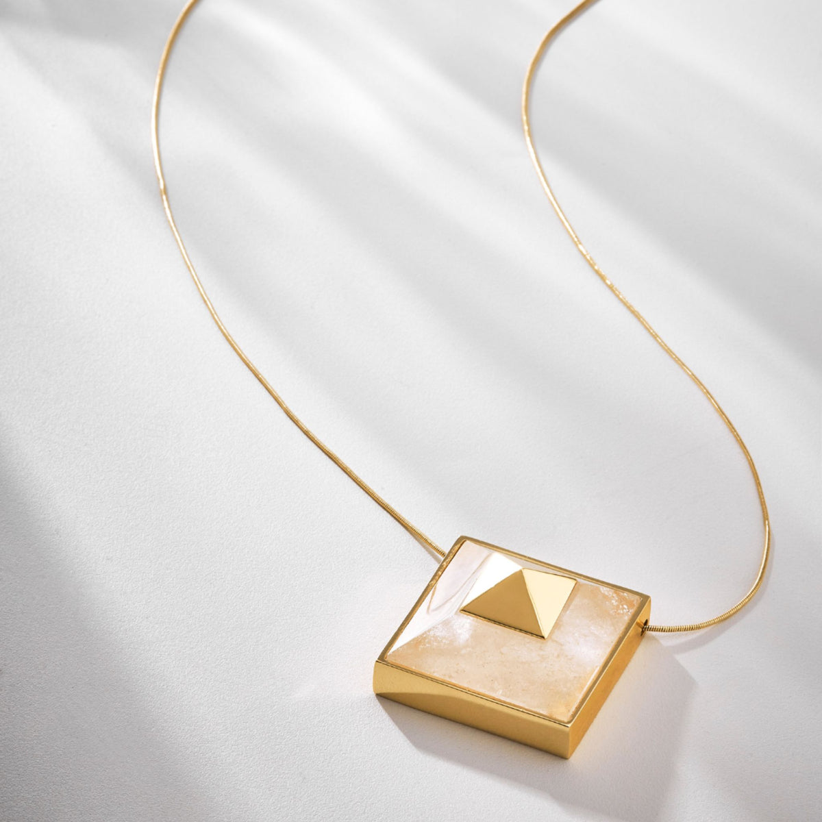 The Gold Vértice L Necklace from ANNA PRATA’s Faces Collection features a thin chain and a square pendant with a raised pyramid design, displayed on a white surface with soft shadows.