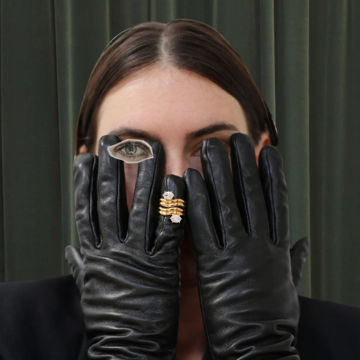 Person wearing black gloves with a gold ring, against a dark curtain background