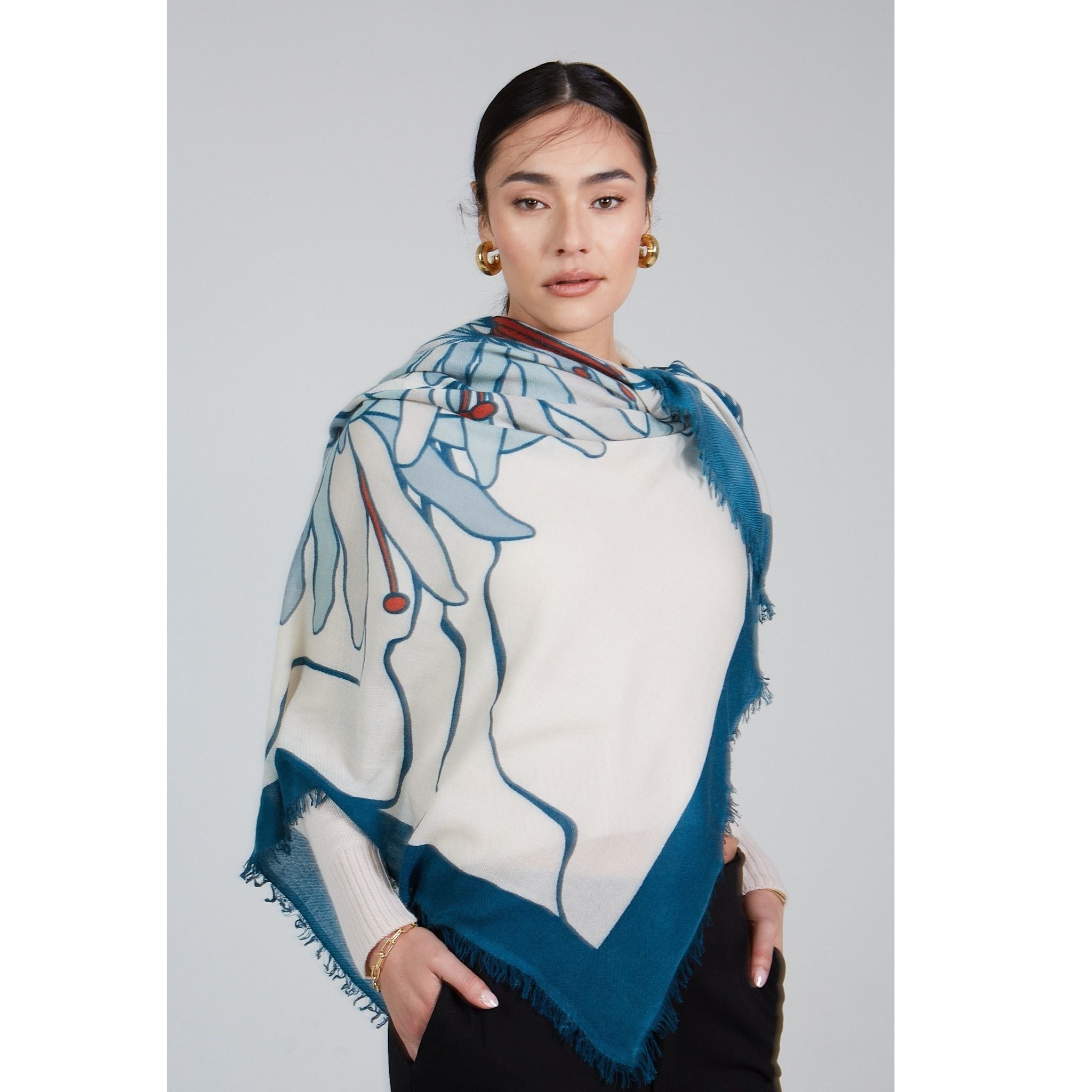 A woman with dark hair tied back wears gold hoop earrings and drapes the OO-TO Olympia Printed Square Scarf—cream and teal cashmere, handcrafted in Nepal with a blue and red abstract leaf pattern—over her shoulders, gazing neutrally to the side.