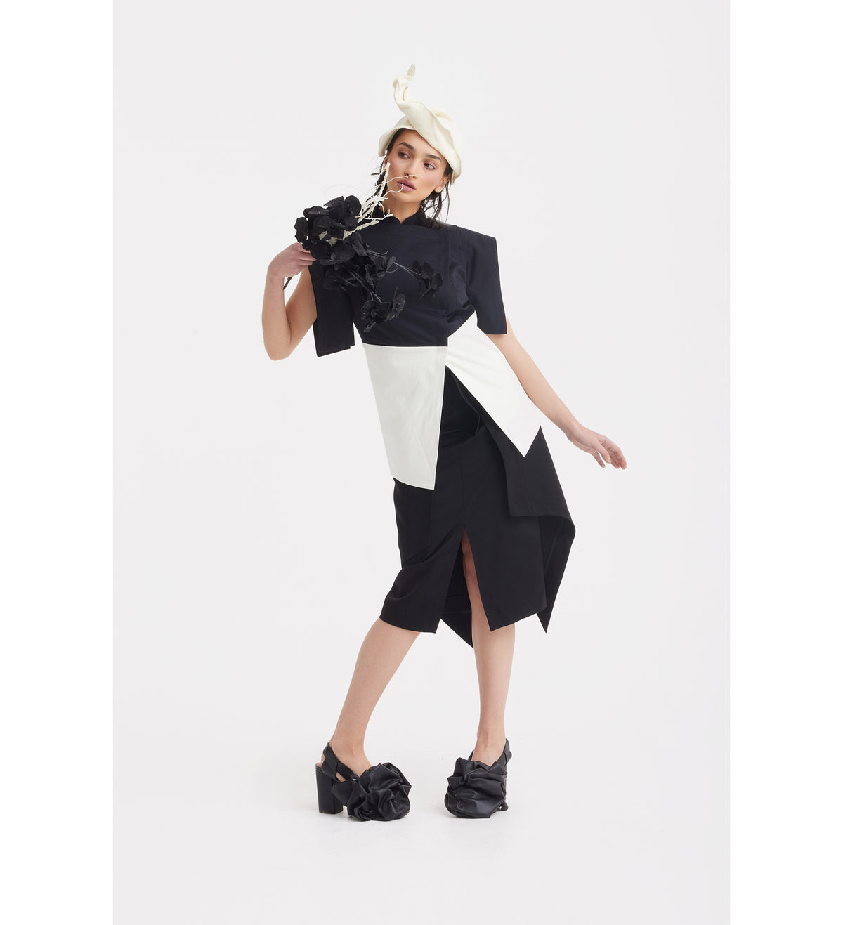 A woman poses in the JULIA ALLERT Asymmetric Colour Block Top - Black &amp; White, styled with black platform shoes featuring large fabric bows and a white hat with black and white accents, against a plain white background.