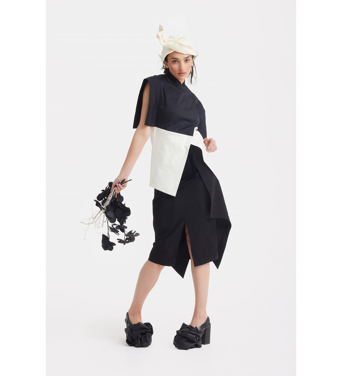 A woman poses in the JULIA ALLERT Asymmetric Colour Block Top in black &amp; white, paired with black platform shoes with bows and a cream beret, holding black flower stems against a plain white background.