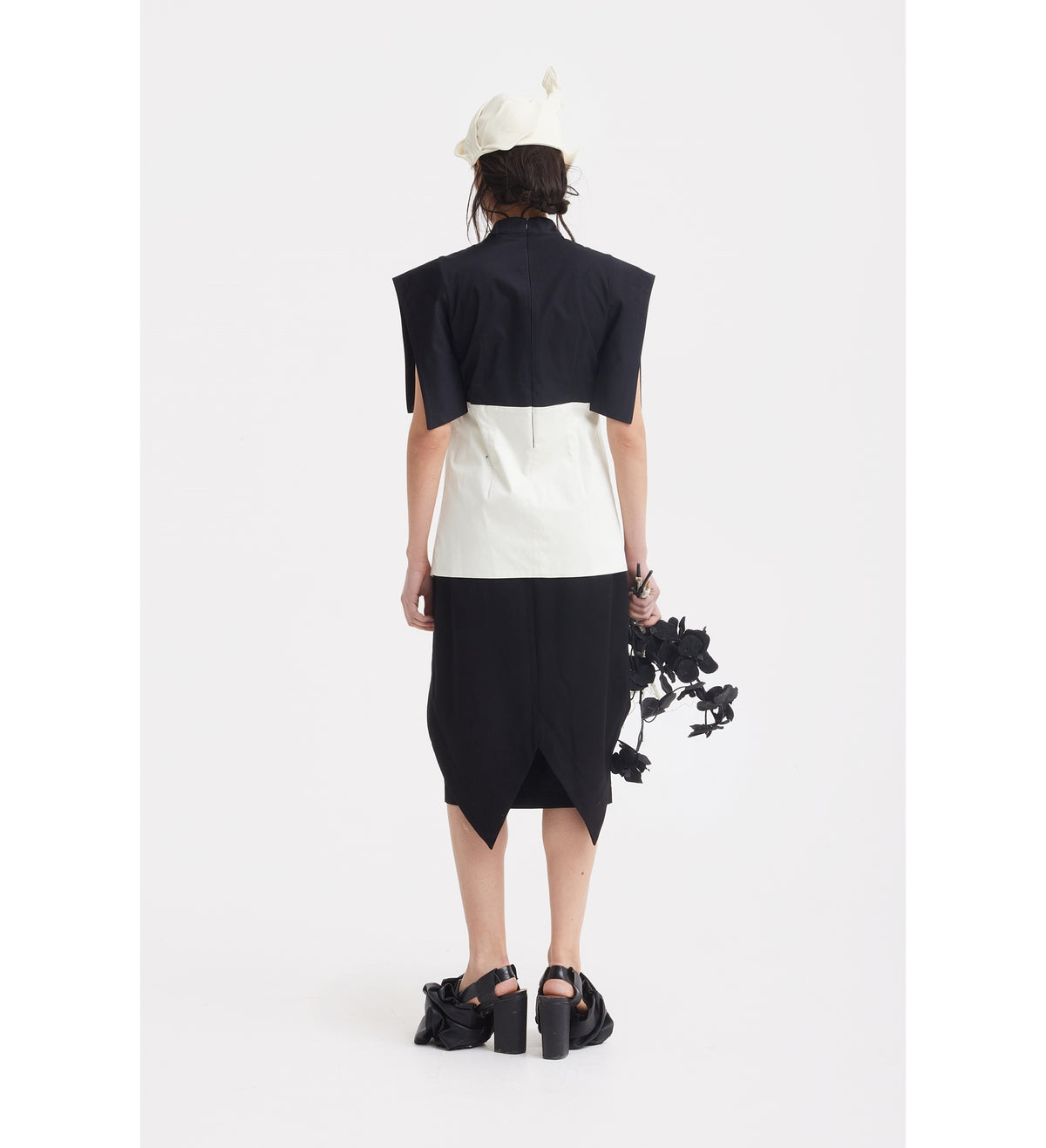 A person faces away, wearing the JULIA ALLERT Asymmetric Colour Block Top in black &amp; white, paired with black platform shoes with bows, a white hat, and holding black artificial flowers against a plain white background.