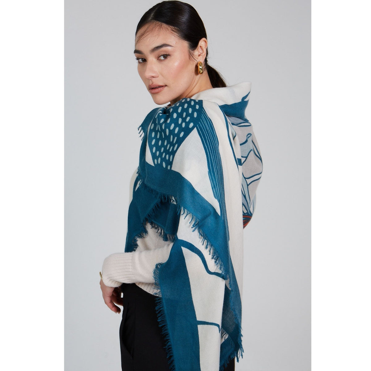 A woman with dark hair pulled back wears gold hoop earrings, a cream sweater, and the OO-TO Olympia Printed Square Scarf in a blue and teal combo, handcrafted in Nepal. She stands against a plain light background, looking over her shoulder.