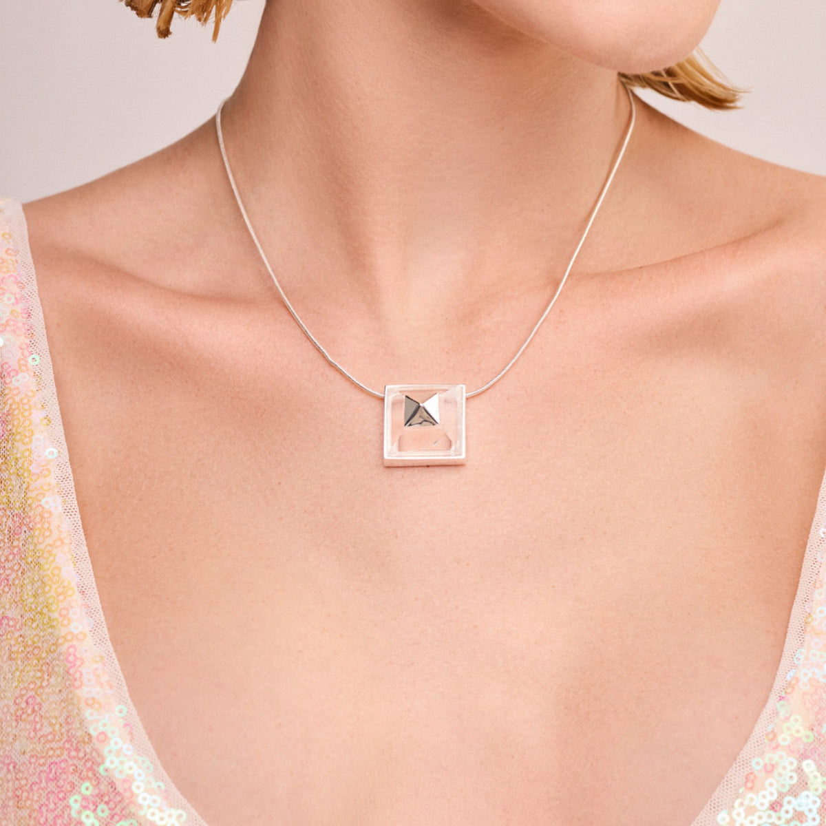 A woman wears the Gold Vértice Necklace from ANNA PRATA's Faces Collection—a delicate gold piece with a square, pyramid-shaped pendant—paired with a light, sequined top that highlights her neck and collarbone.