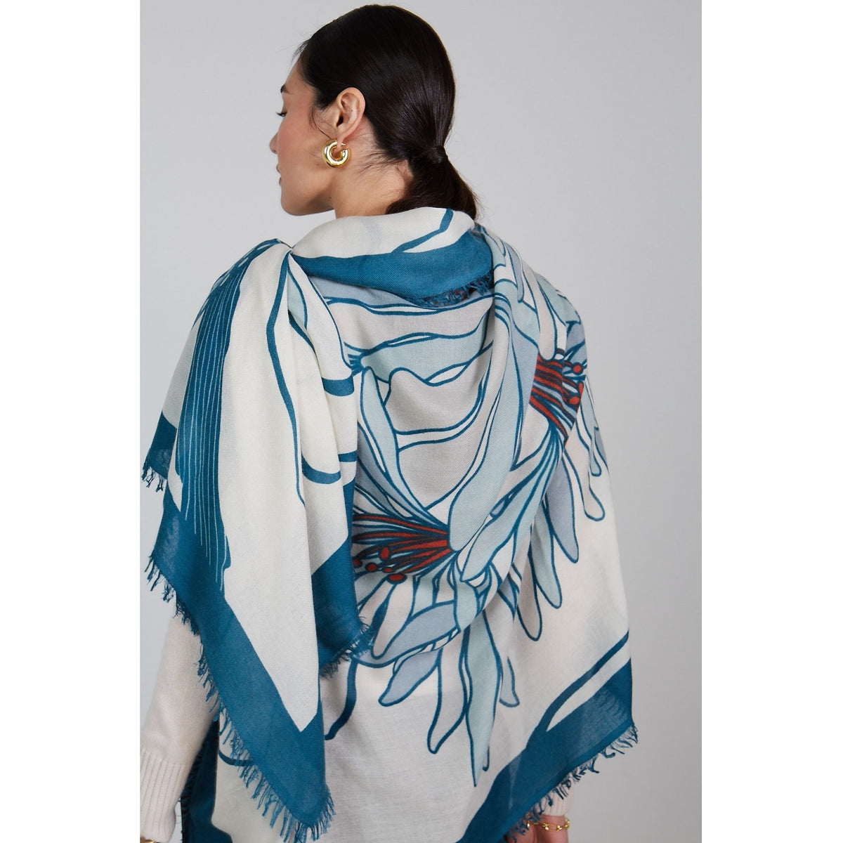 A woman with dark hair in a low ponytail wears gold hoop earrings and the OO-TO Olympia Printed Square Scarf, featuring cream cashmere with blue and red floral patterns, handcrafted in Nepal, draped over her shoulders as she faces away.