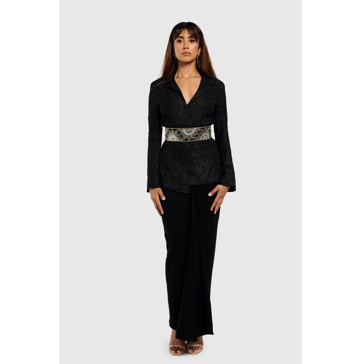 A woman faces forward in the INTERNATIONAL APPEAL Black Paisley Textured Blazer (PR Sample) with a deep neckline, waist belt, and long black skirt. She has straight brown hair, open-toed heels, and is set against a plain background.