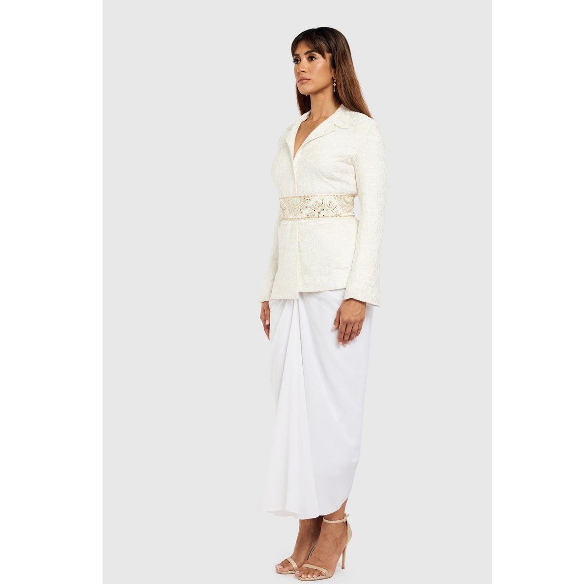 A woman stands against a plain background wearing the White Paisley Textured Blazer by INTERNATIONAL APPEAL, styled with a white draped skirt and nude heels. She looks forward, arms relaxed, embodying modern luxury.