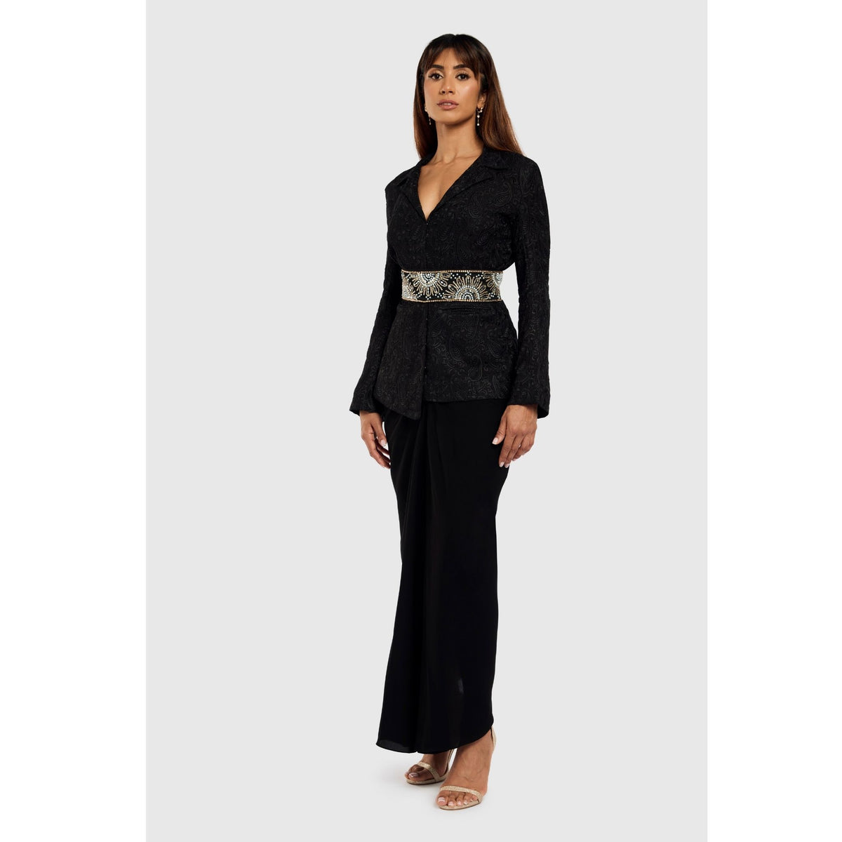 A woman with long brown hair stands confidently against a light background, wearing the INTERNATIONAL APPEAL Black Paisley Textured Blazer (PR Sample) styled with an embellished waist belt and open-toe heels.