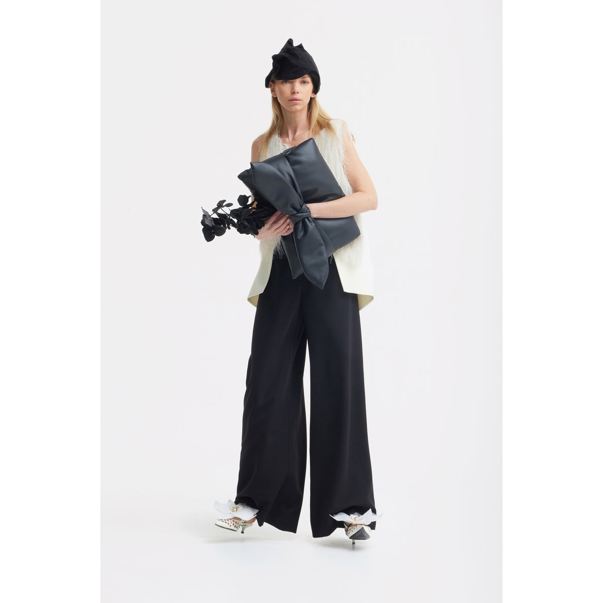 A woman stands against a white background wearing a black beret, sleeveless white top, JULIA ALLERT Statement Waist Wide-Leg Trousers in black, and white shoes. She holds a large black bow and a bouquet of black flowers.