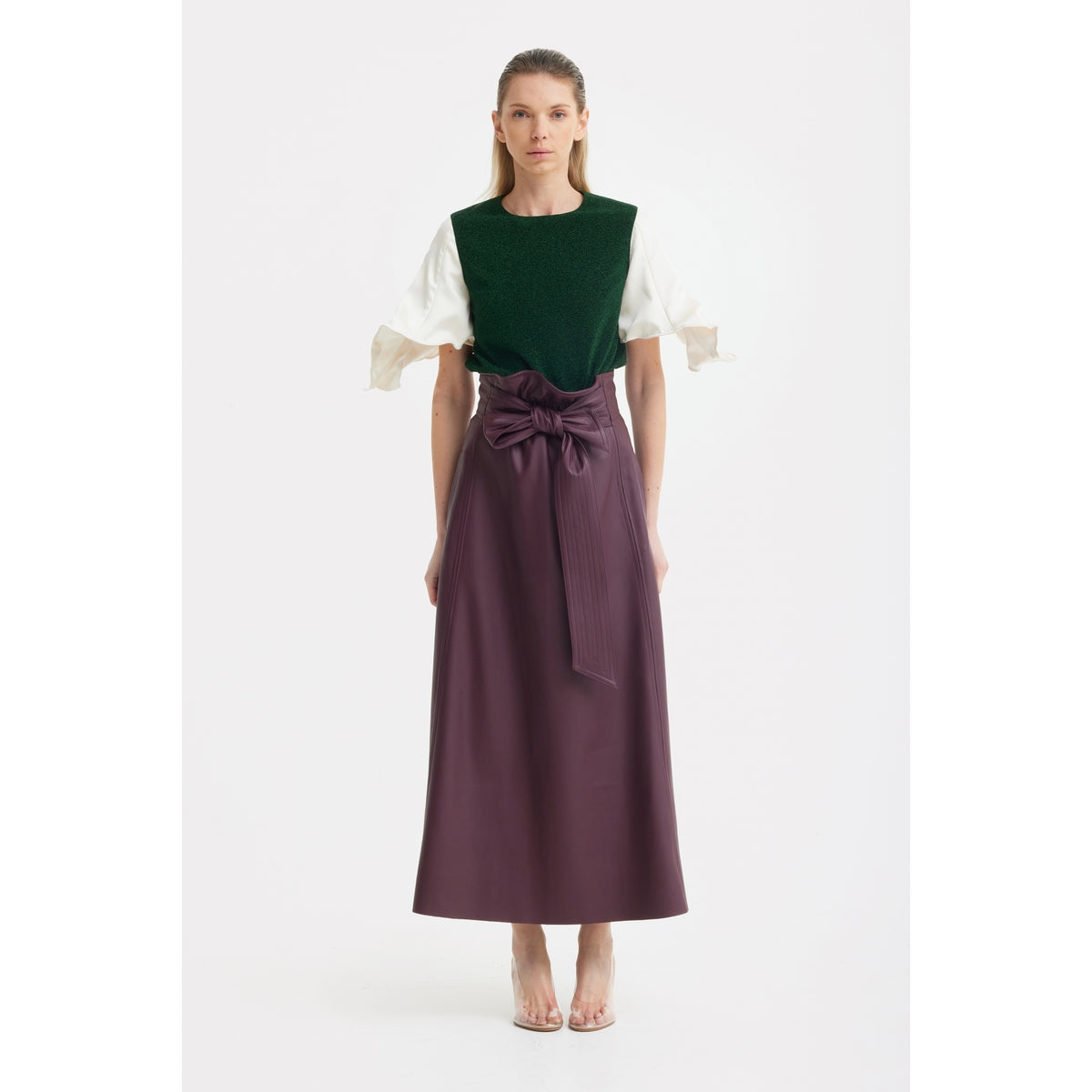 A woman faces forward, wearing a JULIA ALLERT Green Lurex Knit Top with adjustable satin sleeves and a high-waisted, ankle-length purple skirt tied with a large bow. She has straight light brown hair and transparent heels.