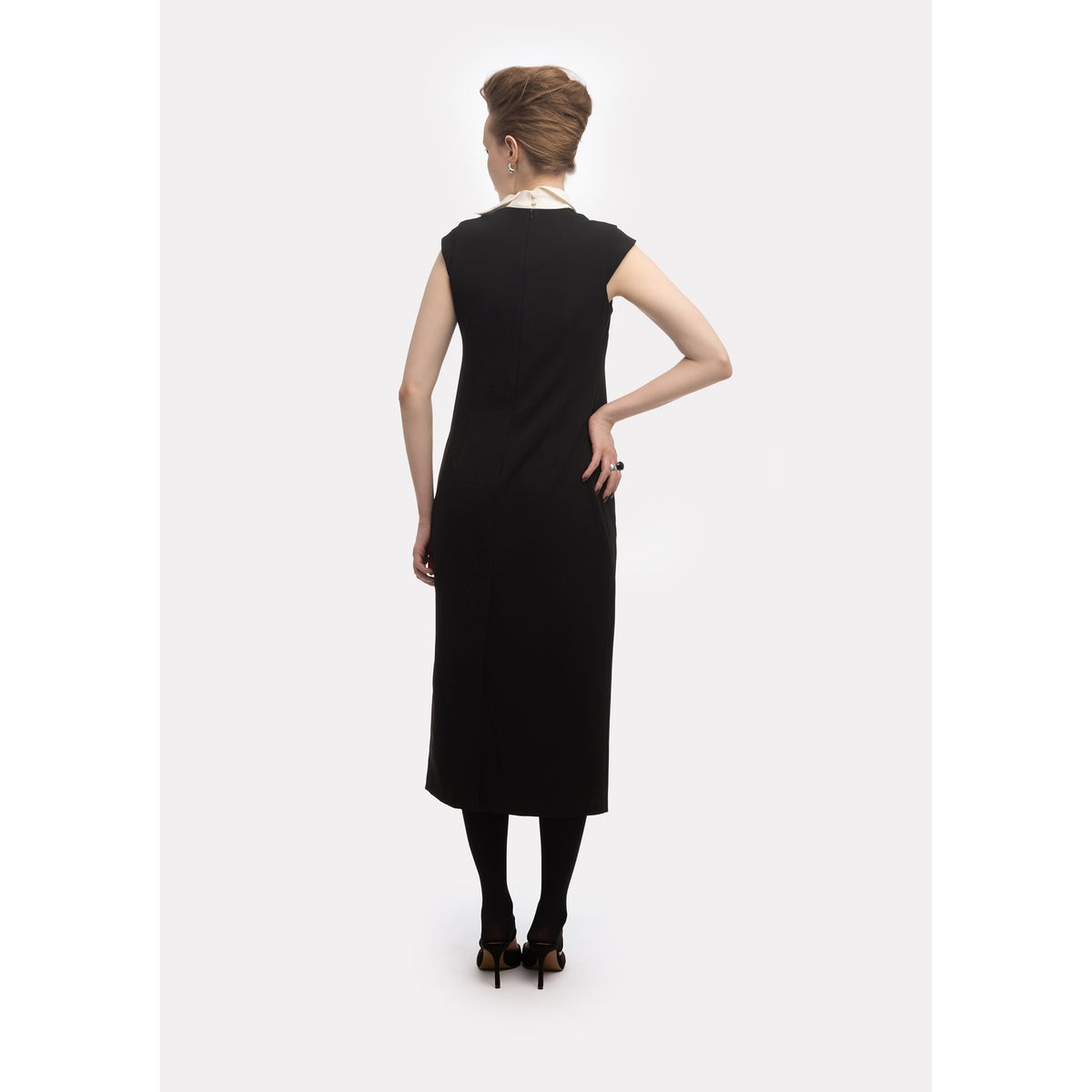 A woman with an updo faces away, wearing the JULIA ALLERT Modern Business Sheath Dress With Decorative Collar in black—sleeveless and knee-length—paired with black tights and heels, set against a plain white background.