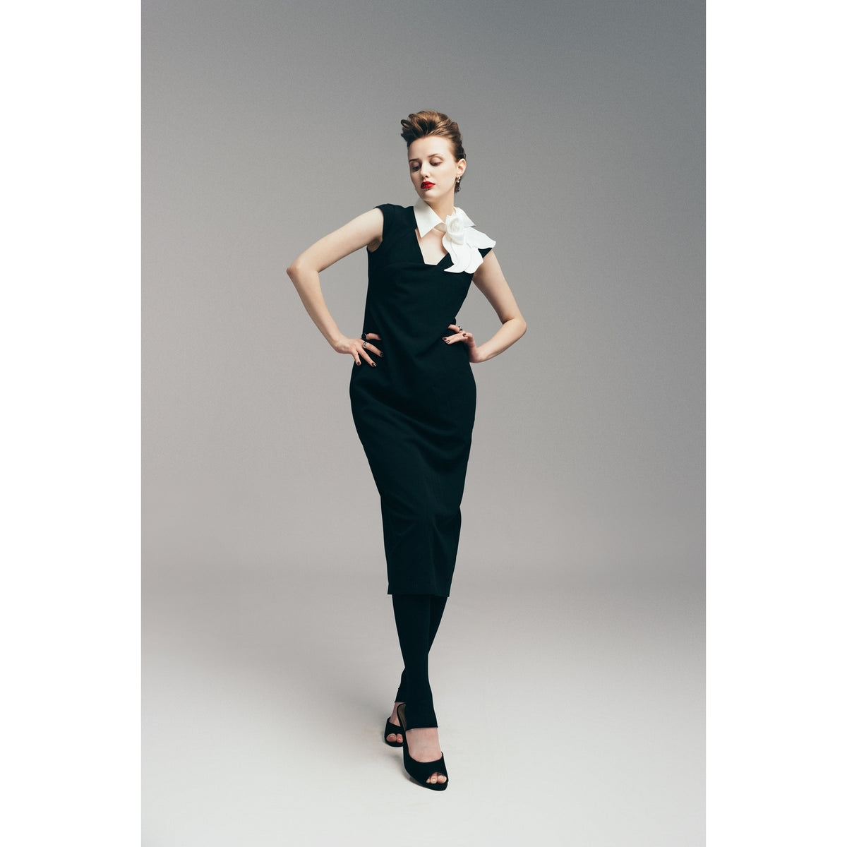 A woman in the JULIA ALLERT Modern Business Sheath Dress With Decorative Collar in black stands against a grey background. She poses with hands on hips, wearing black heels and her eyes closed.