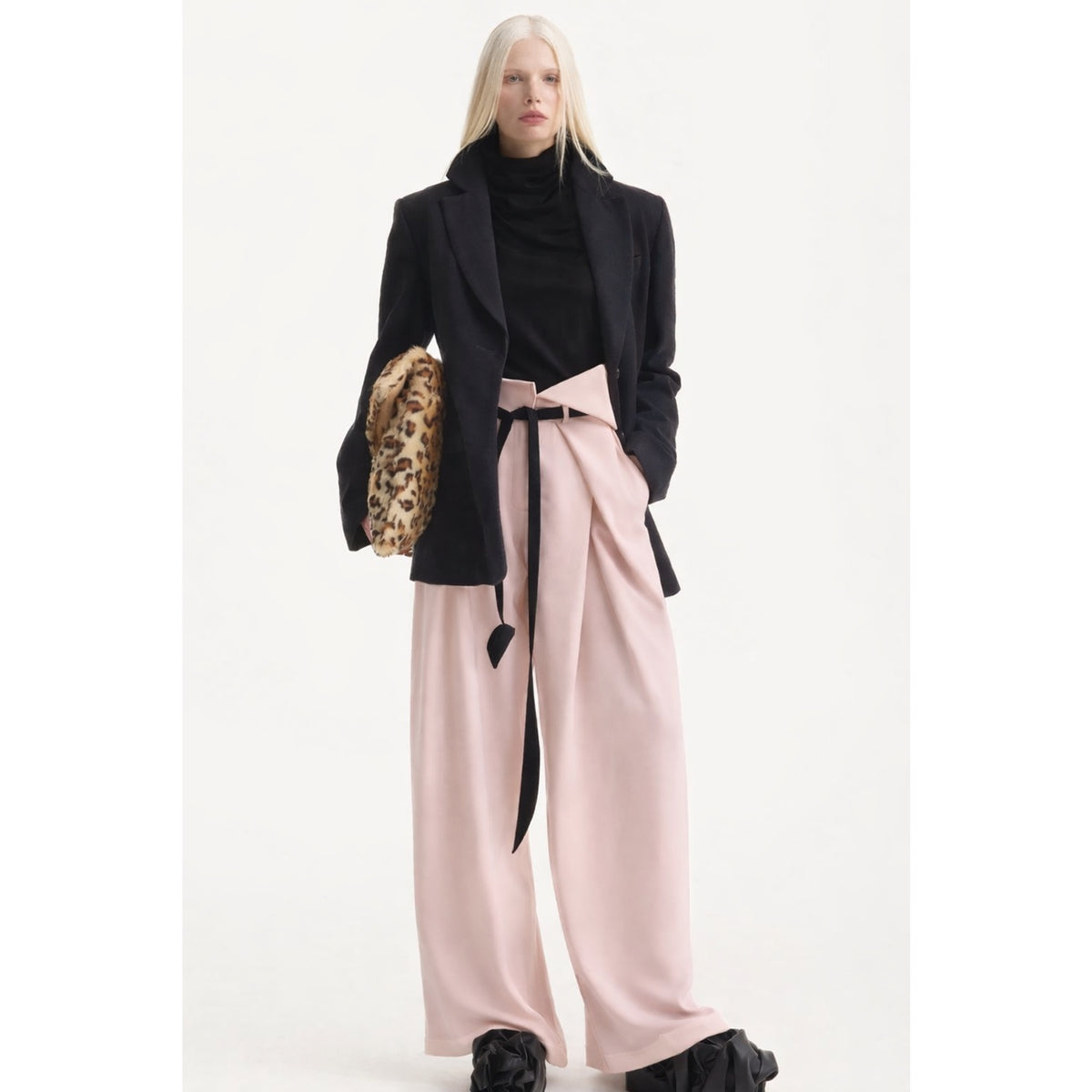 A woman with long blonde hair wears a black turtleneck, blazer, and JULIA ALLERT’s Fashion Forward Wide-Leg Pants &amp; Contrast Belt in Pale Pink. She stands against a plain white background, holding a leopard print item.