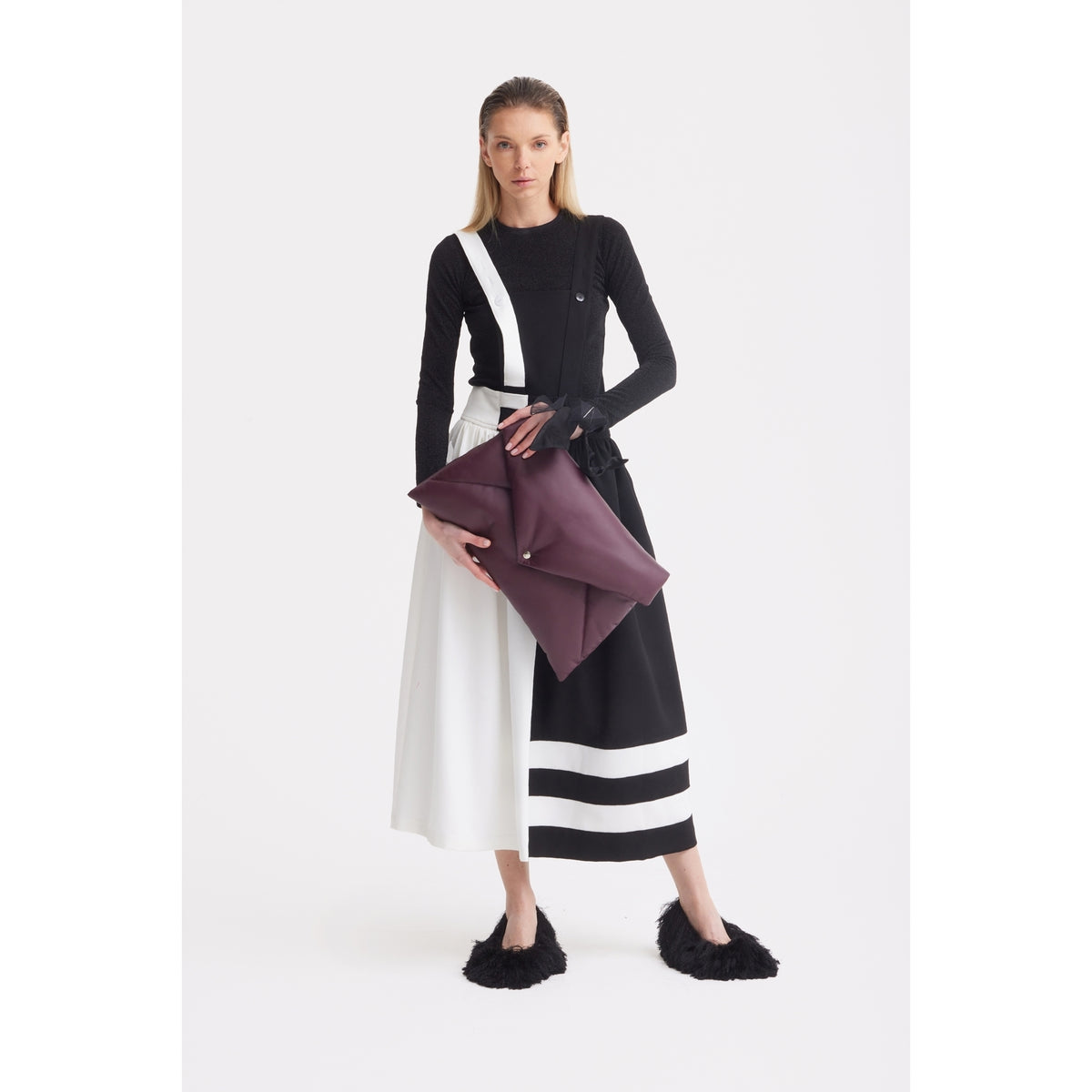 A woman stands against a white background wearing JULIA ALLERT's Midi Apron Dress With Adjustable Straps in Black Ivory, paired with a long-sleeve black top, black furry slippers, and holding a large maroon clutch bag.