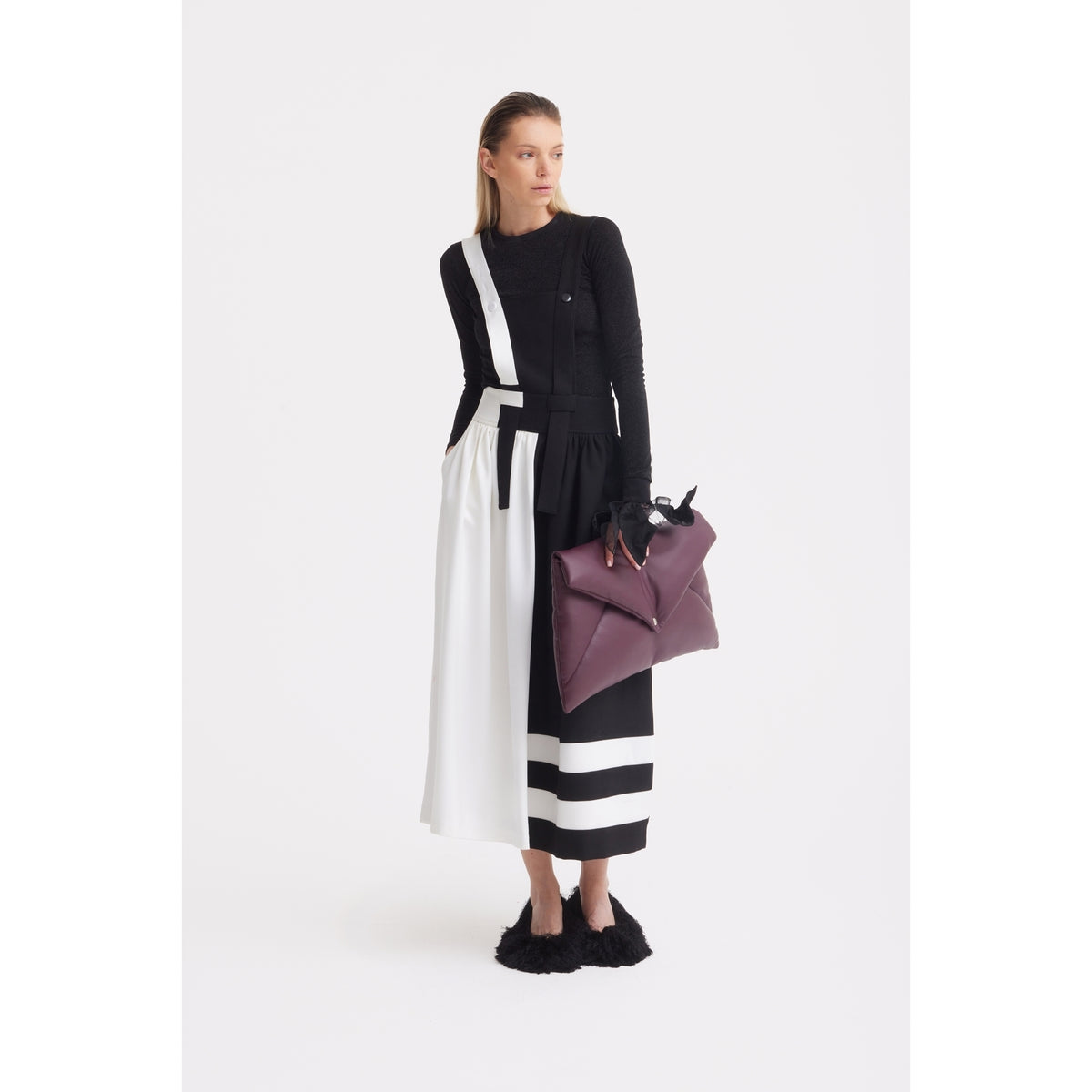 A woman with straight hair stands against a white background, wearing the JULIA ALLERT Midi Apron Dress With Adjustable Straps - Black Ivory, geometric patterns, black textured shoes, and holding a large purple handbag while looking slightly to the side.