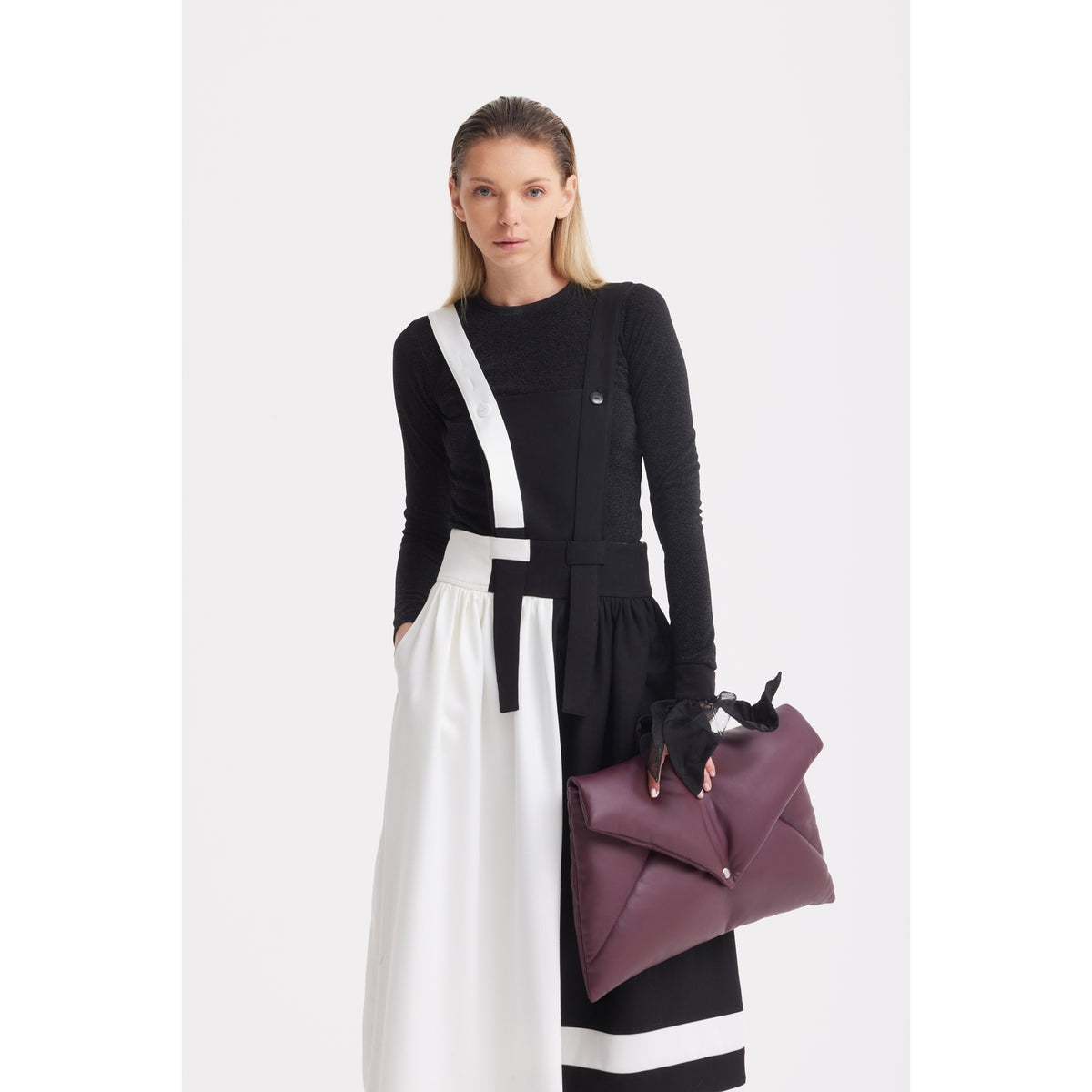 A woman stands against a white background wearing the JULIA ALLERT Midi Apron Dress With Adjustable Straps - Black Ivory over a black long-sleeve top, holding a large purple envelope-style clutch.