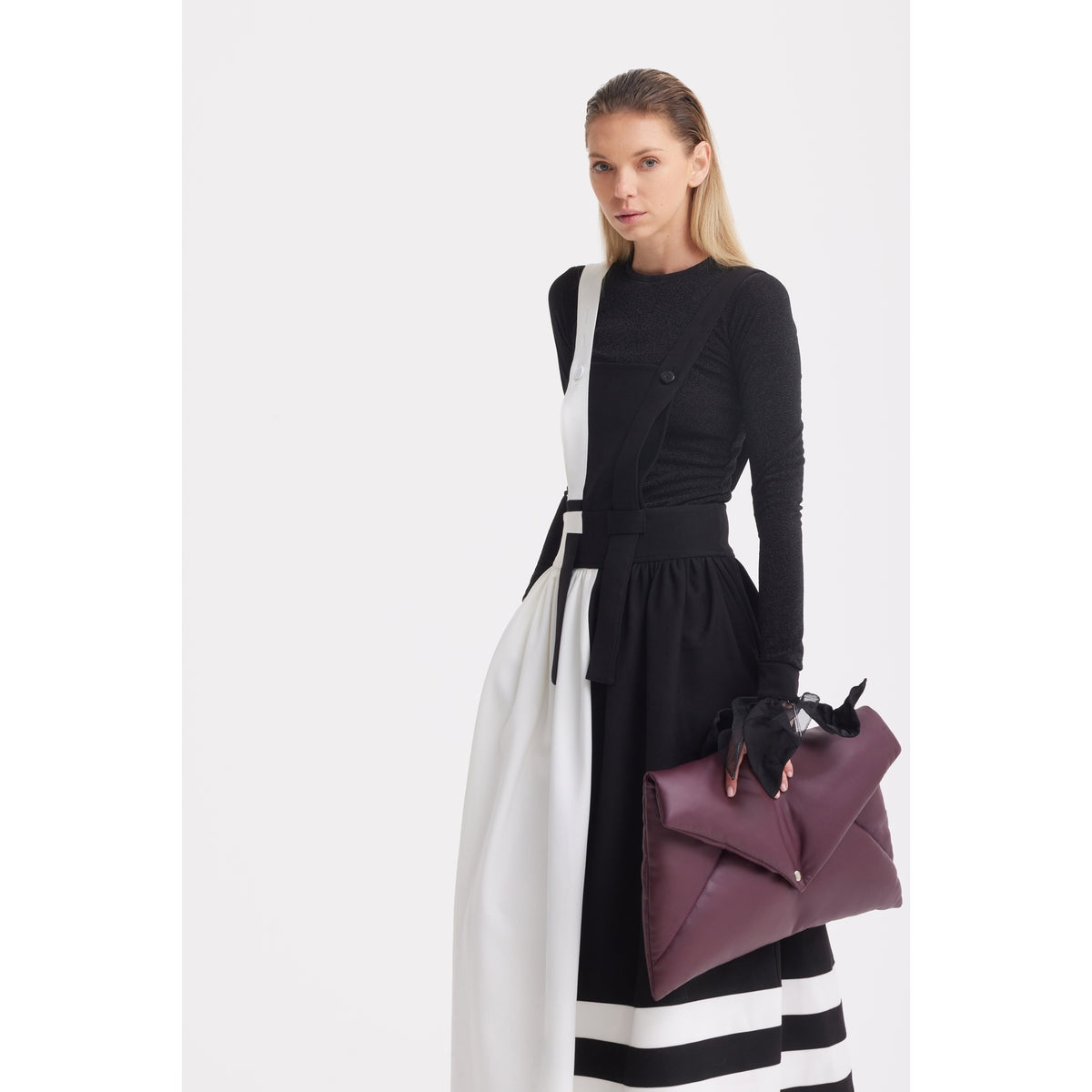 A blonde model wears the JULIA ALLERT Midi Apron Dress With Adjustable Straps in Black Ivory, paired with a burgundy quilted leather clutch featuring a black bow detail.