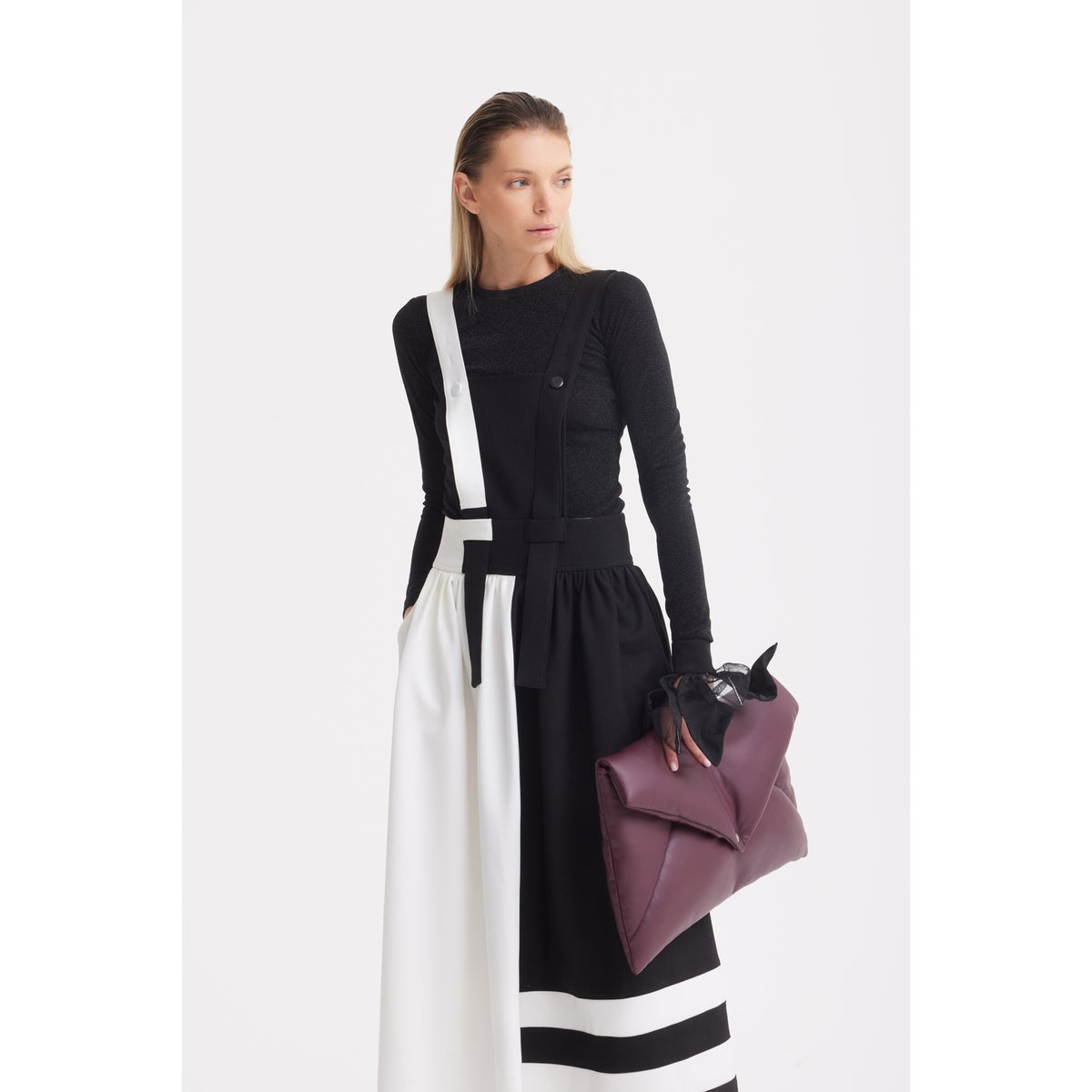 A woman with long blonde hair wears a black long-sleeve top and the JULIA ALLERT Midi Apron Dress With Adjustable Straps - Black Ivory, holding a large plum clutch as she looks left against a plain white background.