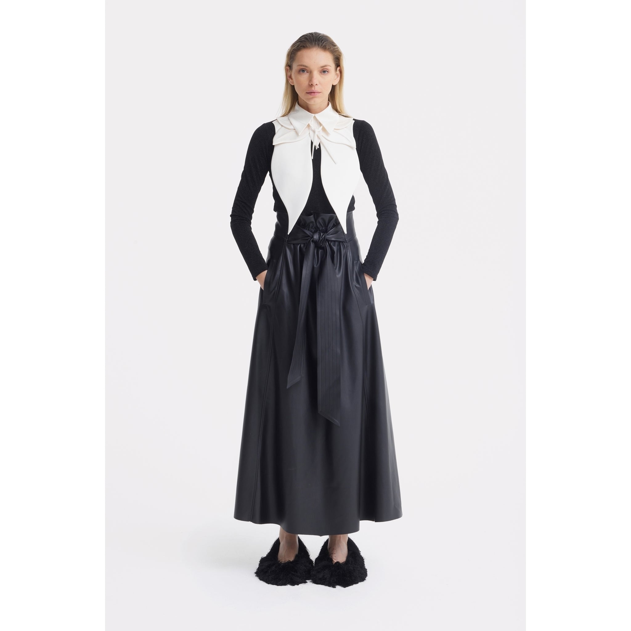A woman stands facing forward in a black long-sleeve top, long belted skirt, and black fuzzy slippers. Her straight hair is loose and her hands are in her pockets. She finishes the look with JULIA ALLERT’s Oversized Satin Bib Collar in Ivory.