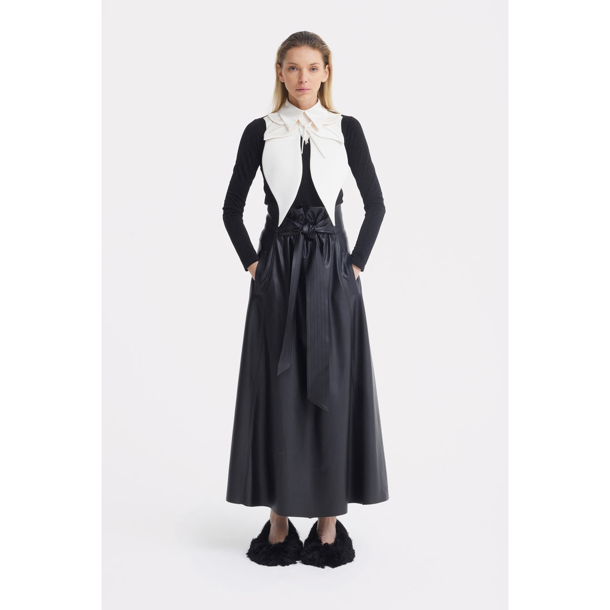 A woman stands facing forward in a black long-sleeve top, long belted skirt, and black fuzzy slippers. Her straight hair is loose and her hands are in her pockets. She finishes the look with JULIA ALLERT’s Oversized Satin Bib Collar in Ivory.
