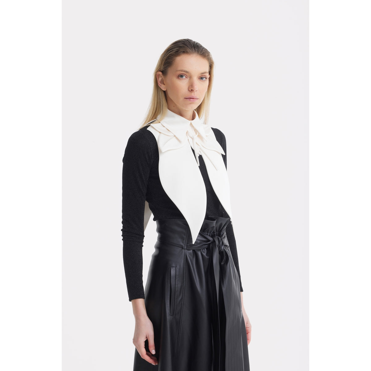 A woman with straight blonde hair wears a black long-sleeve top, a high-waisted black leather skirt with a tie belt, and the JULIA ALLERT Oversized Satin Bib Collar in ivory, standing against a plain white background.