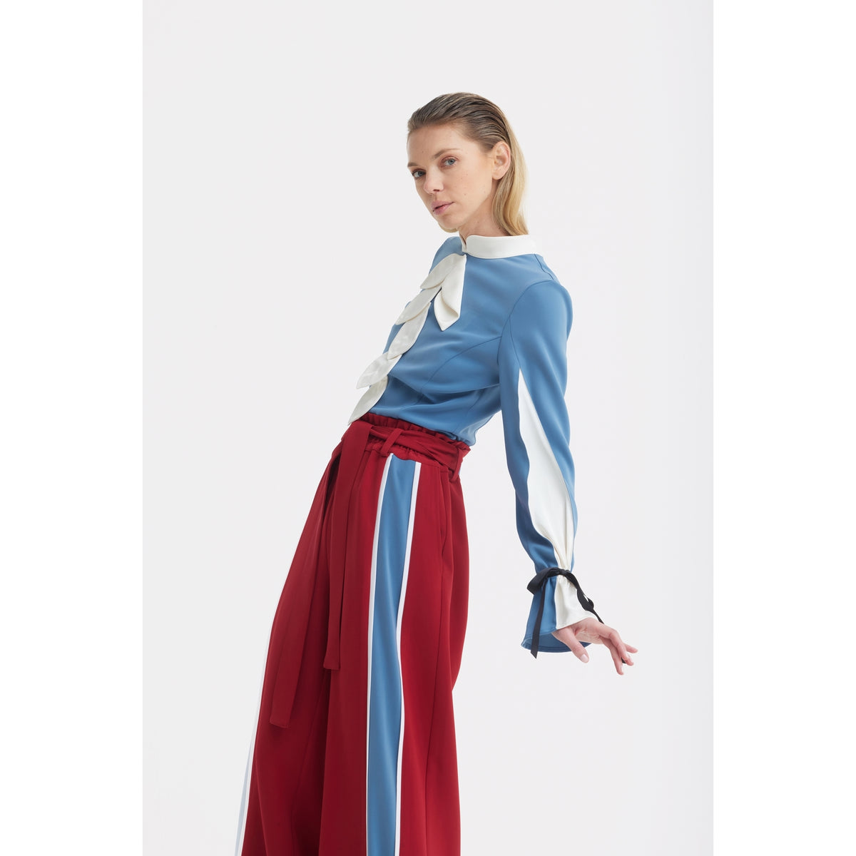 A person poses against a white background wearing the JULIA ALLERT Elegant Fitted Blouse - Blue, featuring white accents and a bow at the collar, paired with high-waisted red luxury suiting pants.