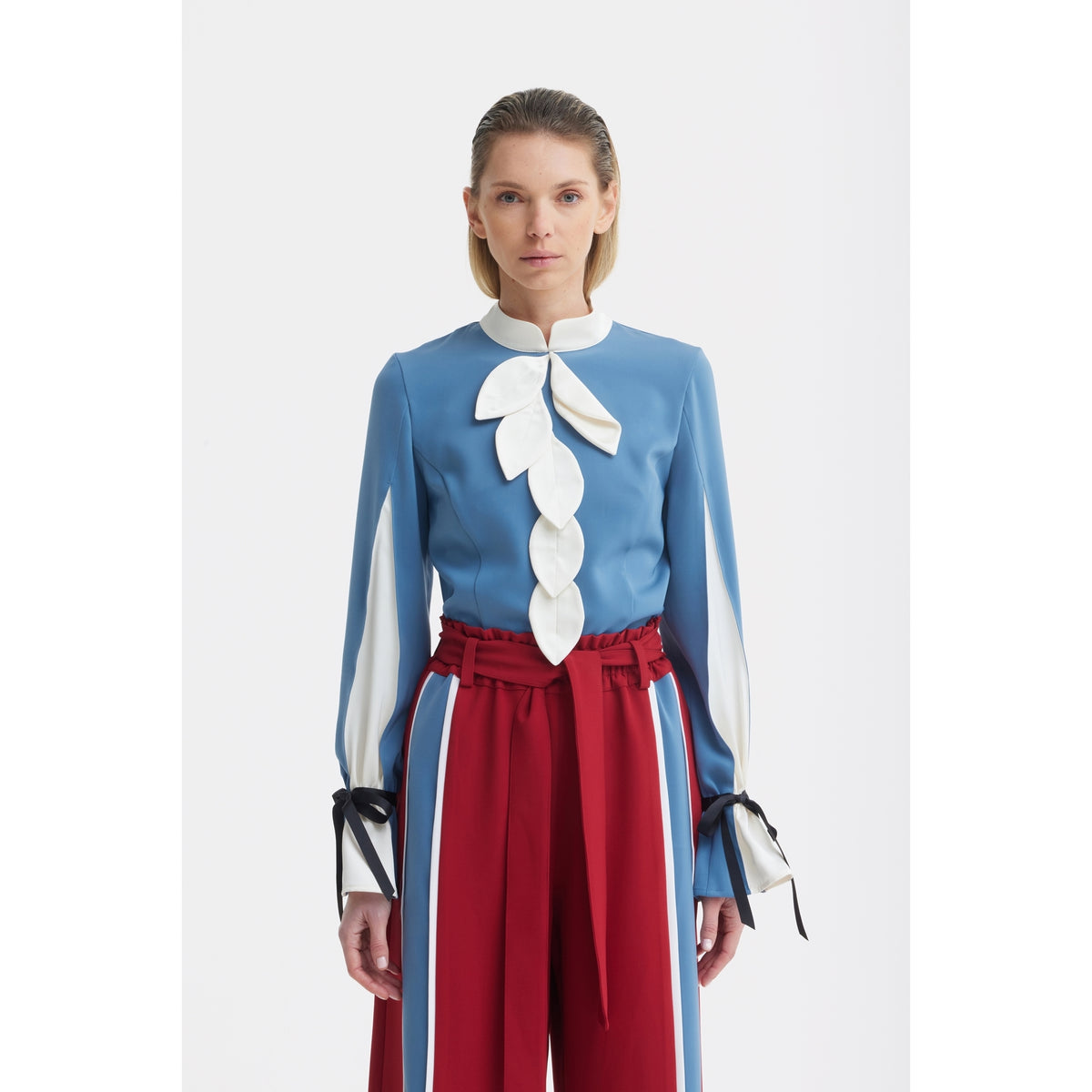 A woman faces forward in JULIA ALLERT's Elegant Fitted Blouse - Blue, detailed with white leaf accents and black ribbon-tied long sleeves, paired with high-waisted red trousers featuring blue and white side stripes.